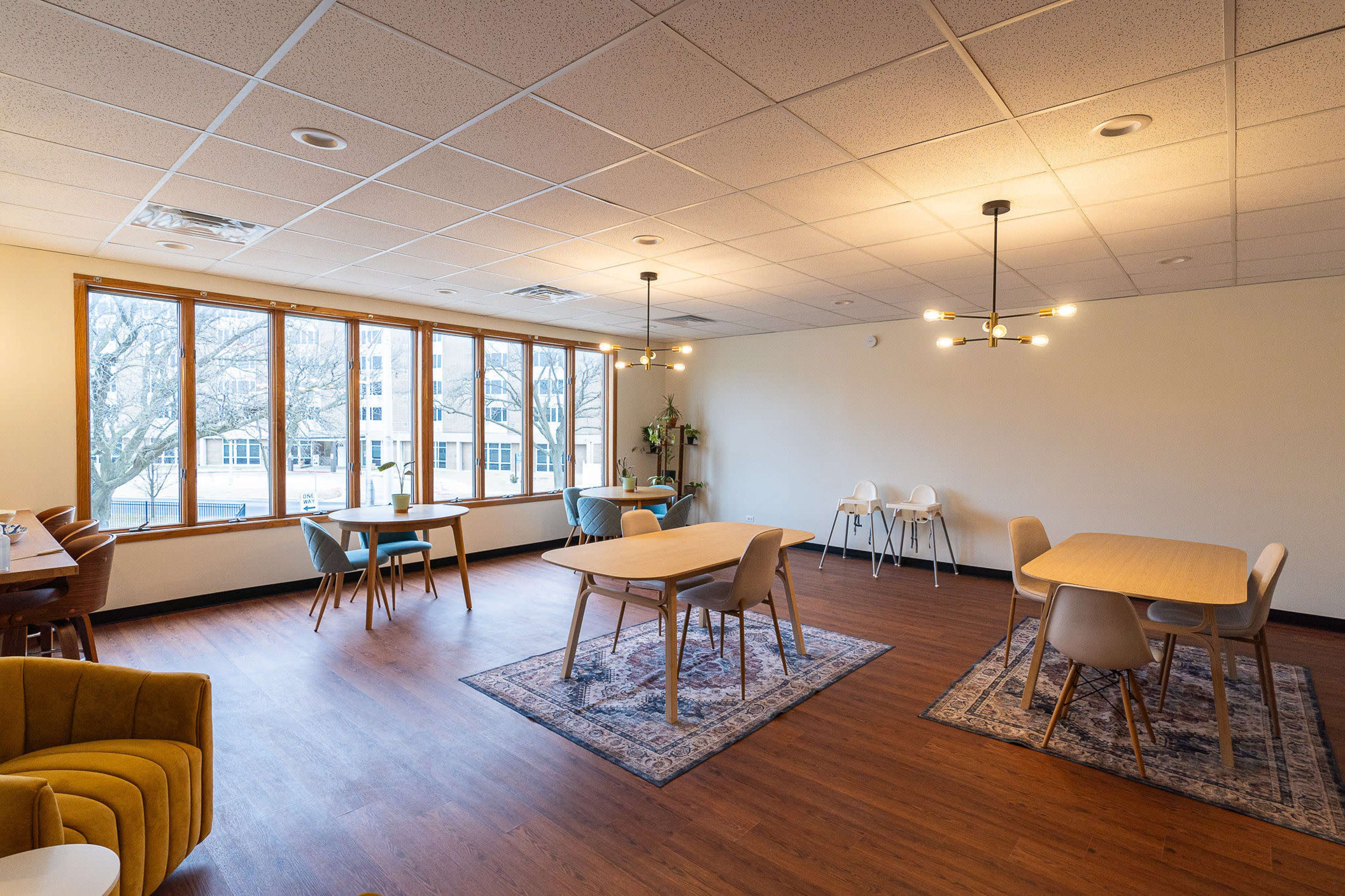 The image shows a spacious, well-lit room furnished with several tables and chairs, featuring large windows that provide views of trees outside.