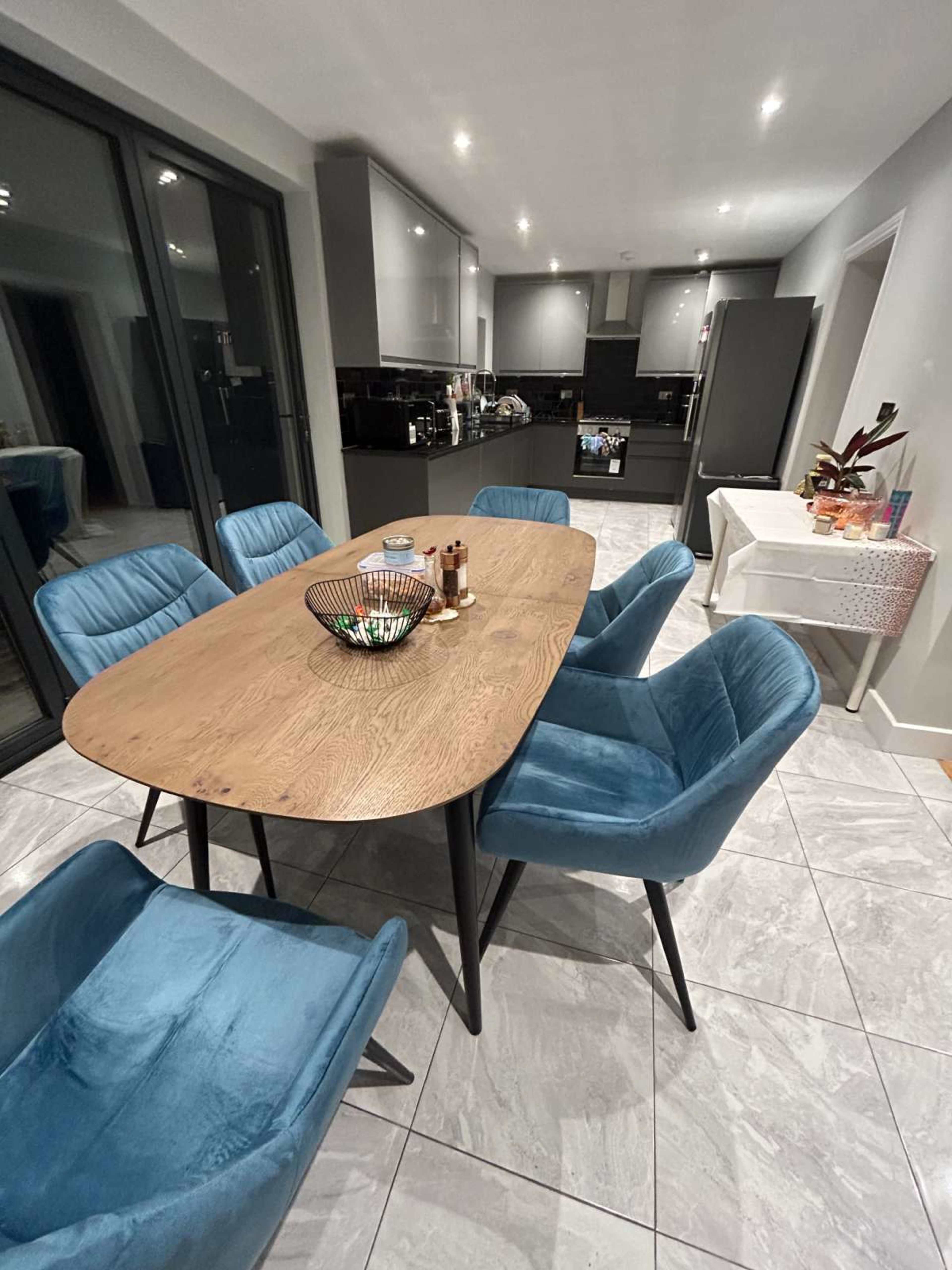 A modern dining area features a wooden oval table surrounded by six blue upholstered chairs, with a side table set up in the corner.