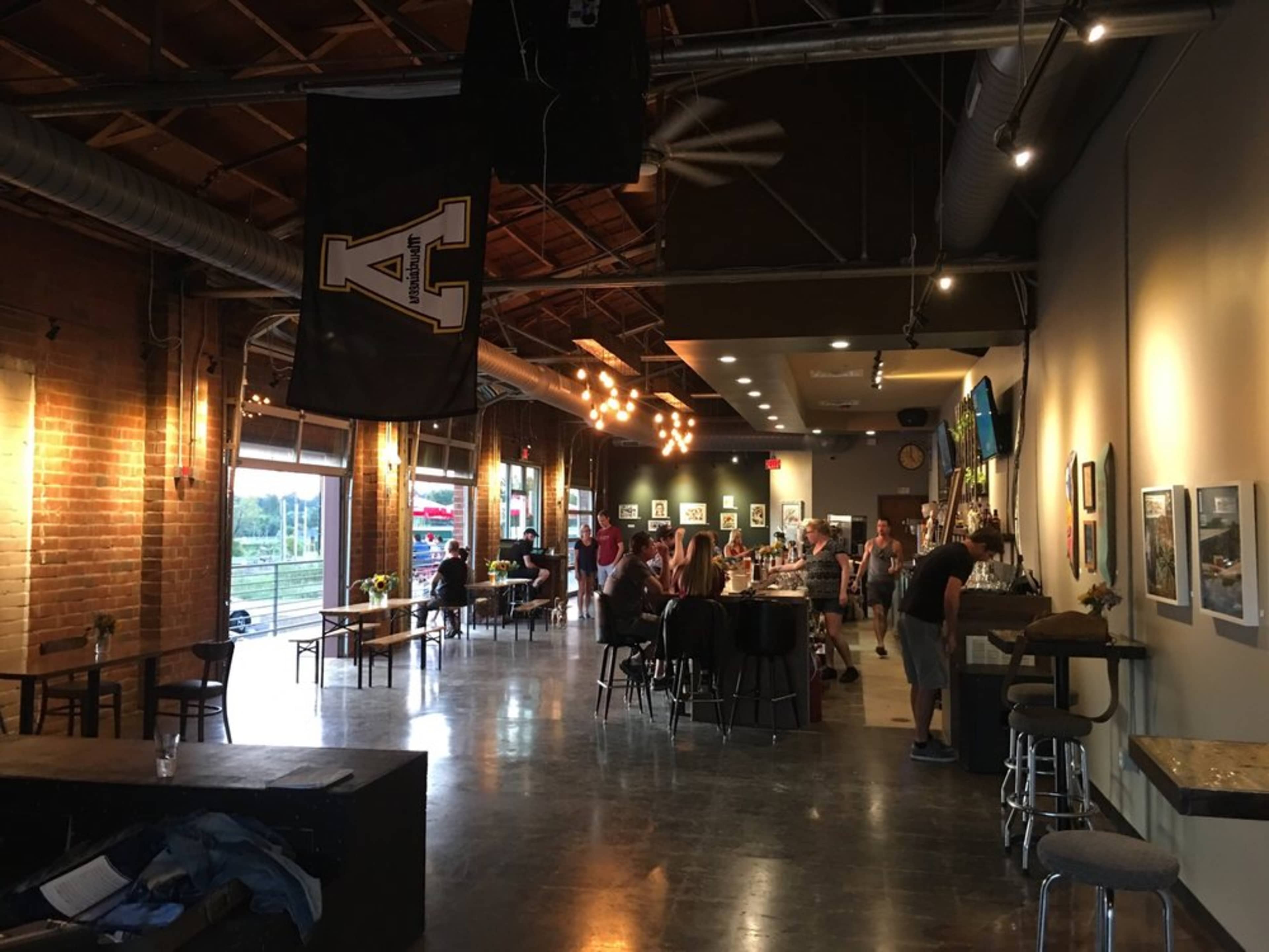 The image shows a spacious, industrial-style café with brick walls, wooden tables, and a few people seated or lining up at the counter.