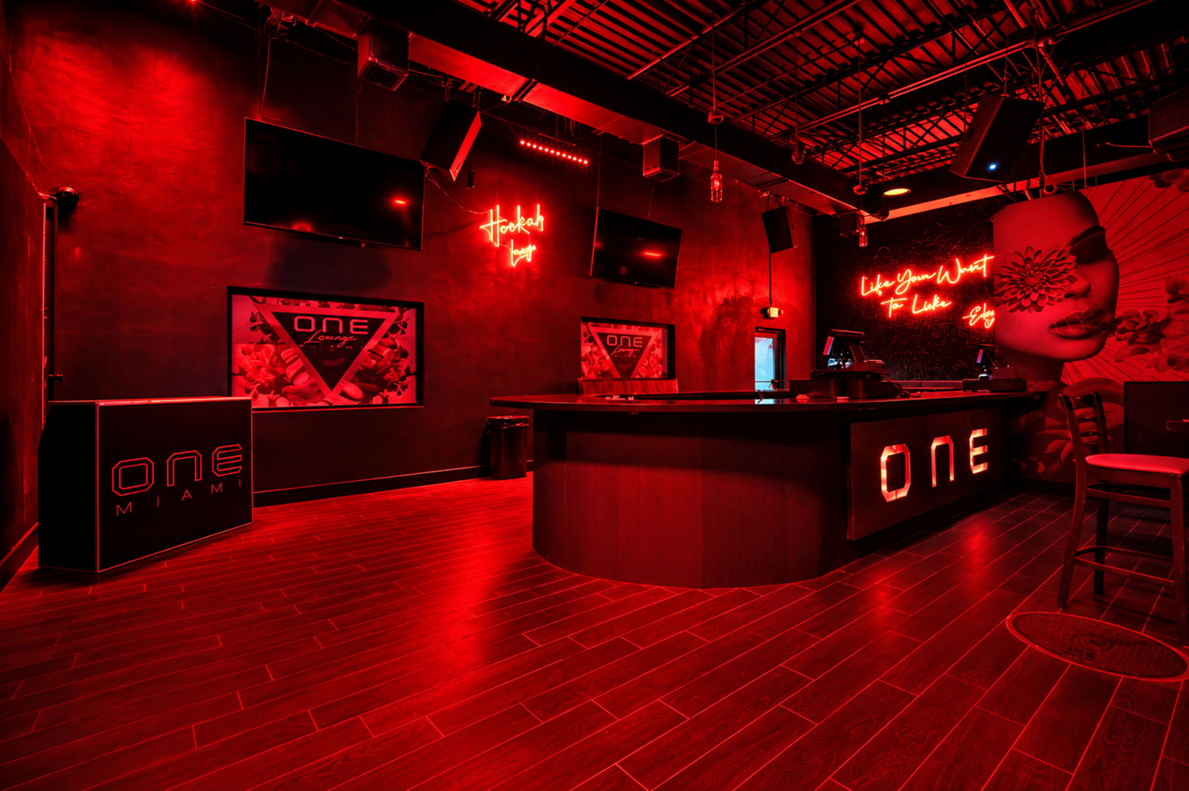 The interior of a dimly lit bar features red walls, a sleek bar area with an illuminated sign reading "ONE," and multiple screens mounted on the walls.
