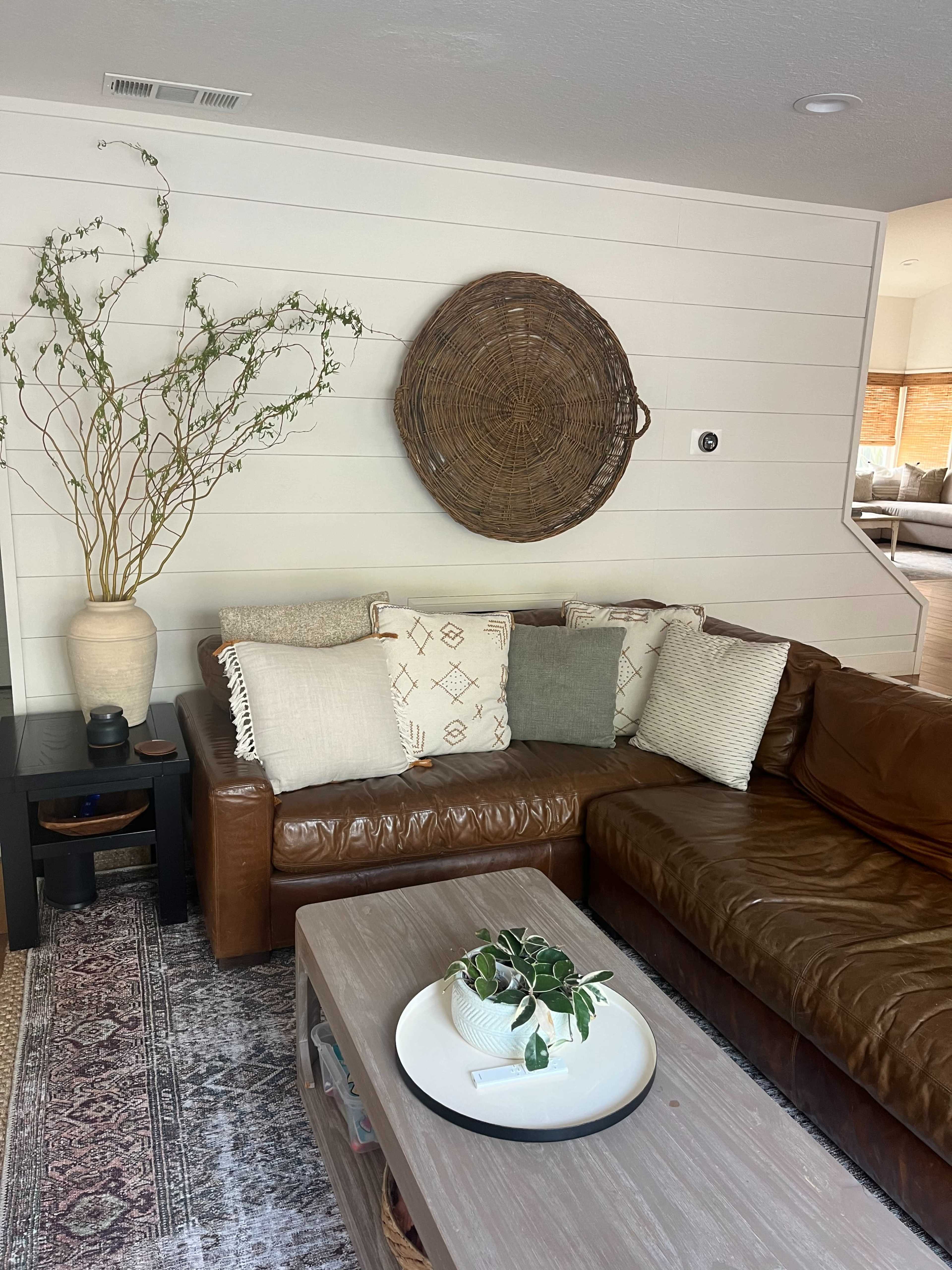 A cozy living room features a brown leather sectional sofa, a wooden coffee table with a decorative plant, and a large woven wall art piece above the seating area.