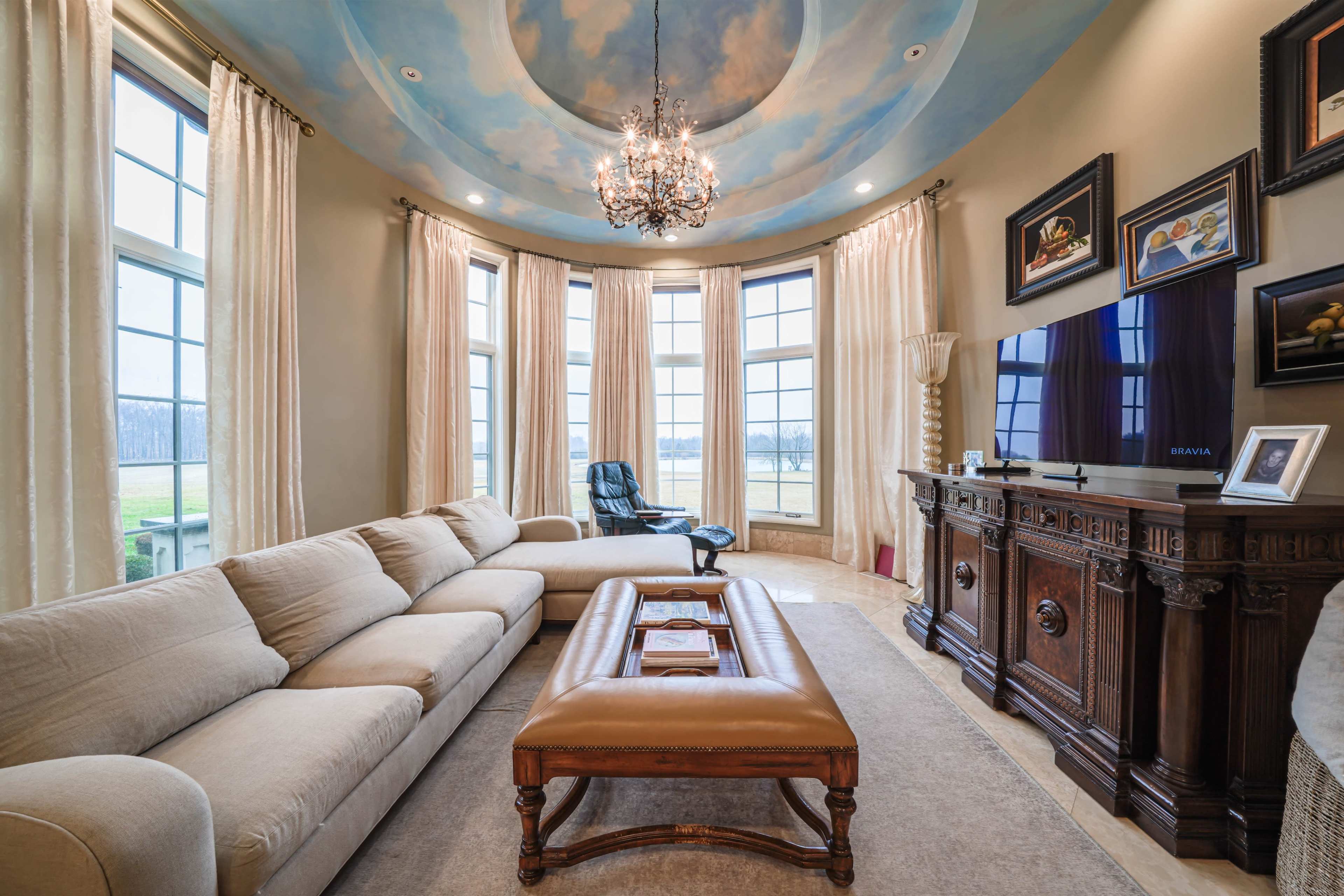 A spacious living room features a large sectional sofa, an ornate wooden cabinet, and a chandelier under a painted ceiling, with large windows offering a view of the outdoors.