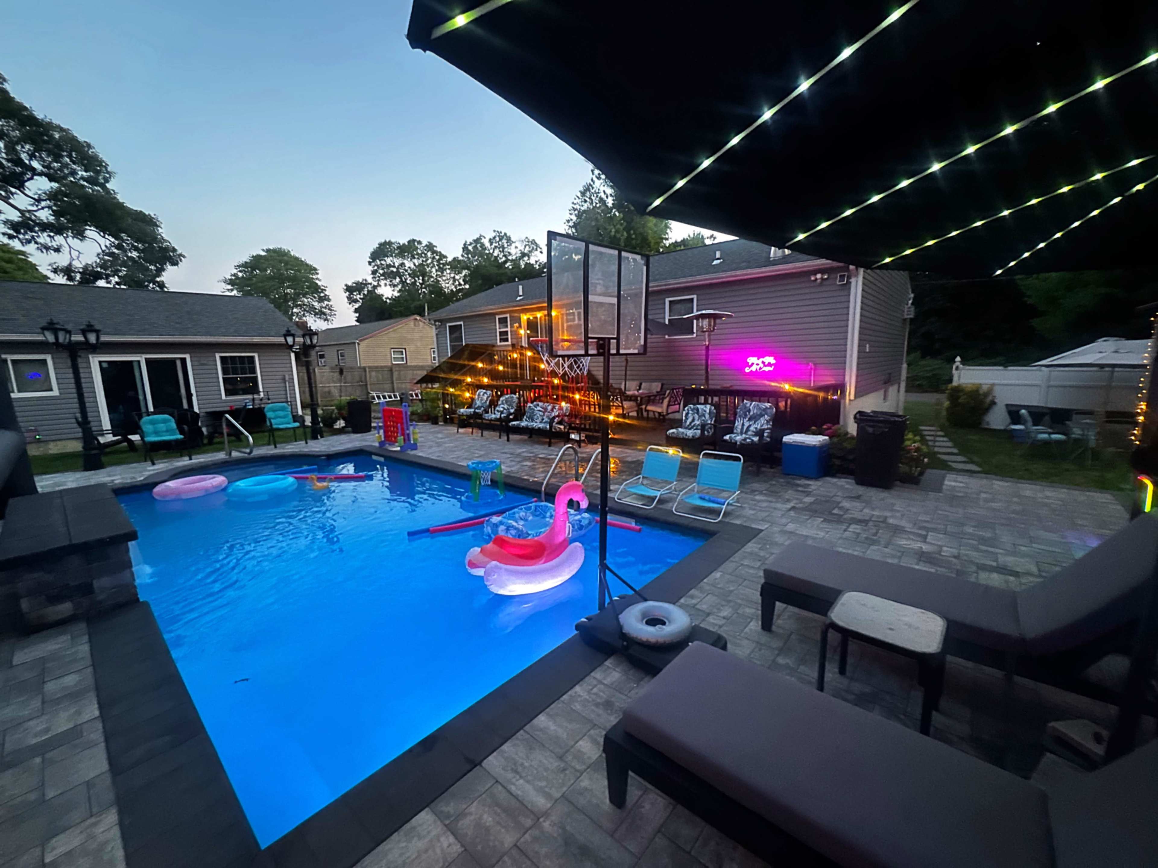 The image shows a backyard pool area with colorful floating accessories, lounge chairs, and string lights decorating a nearby structure as evening approaches.