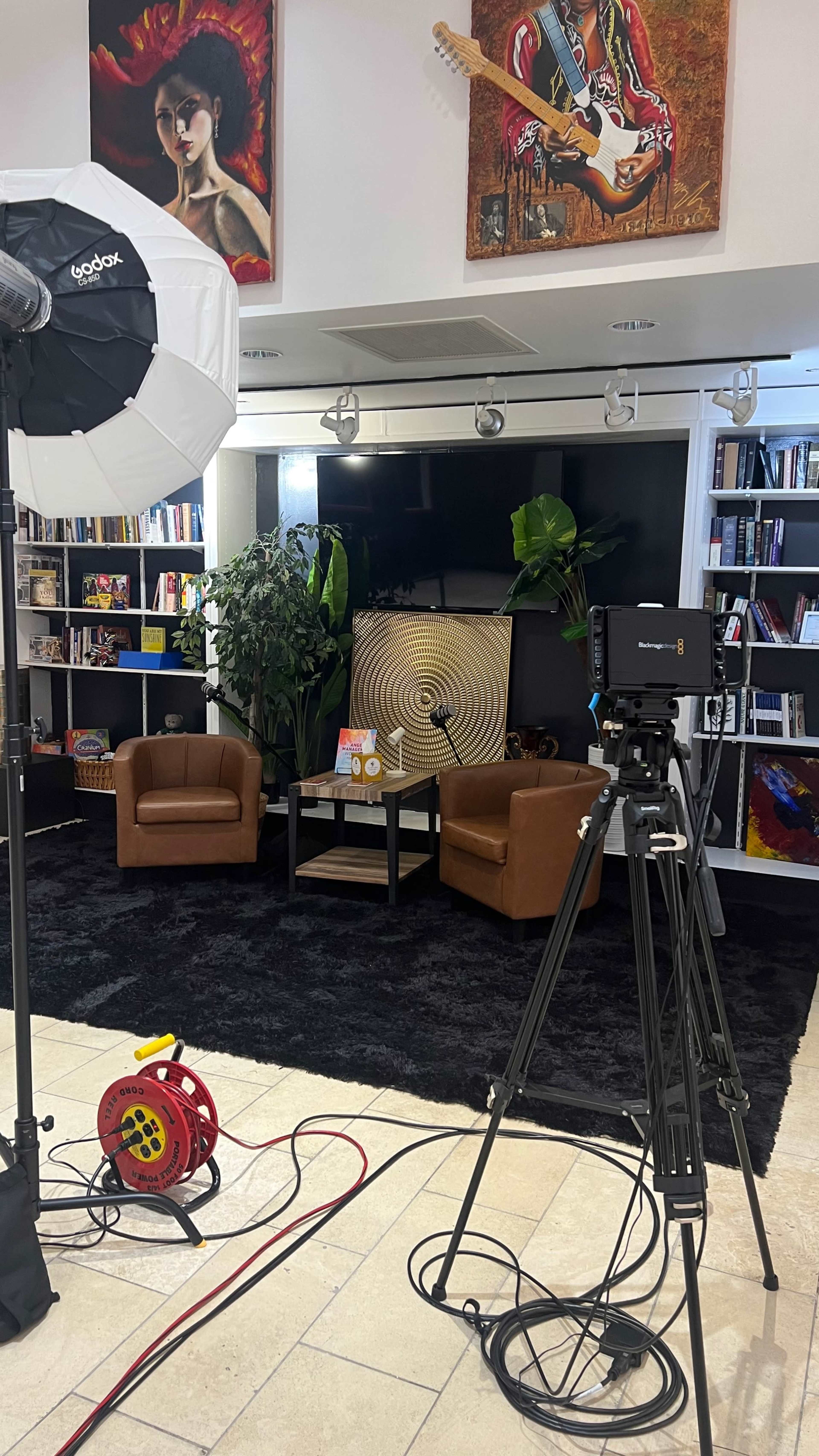 The image shows a modern studio setup featuring two brown armchairs, a black rug, a backdrop display with a circular pattern, and a camera facing the seating area, surrounded by bookshelves and artwork on the walls.