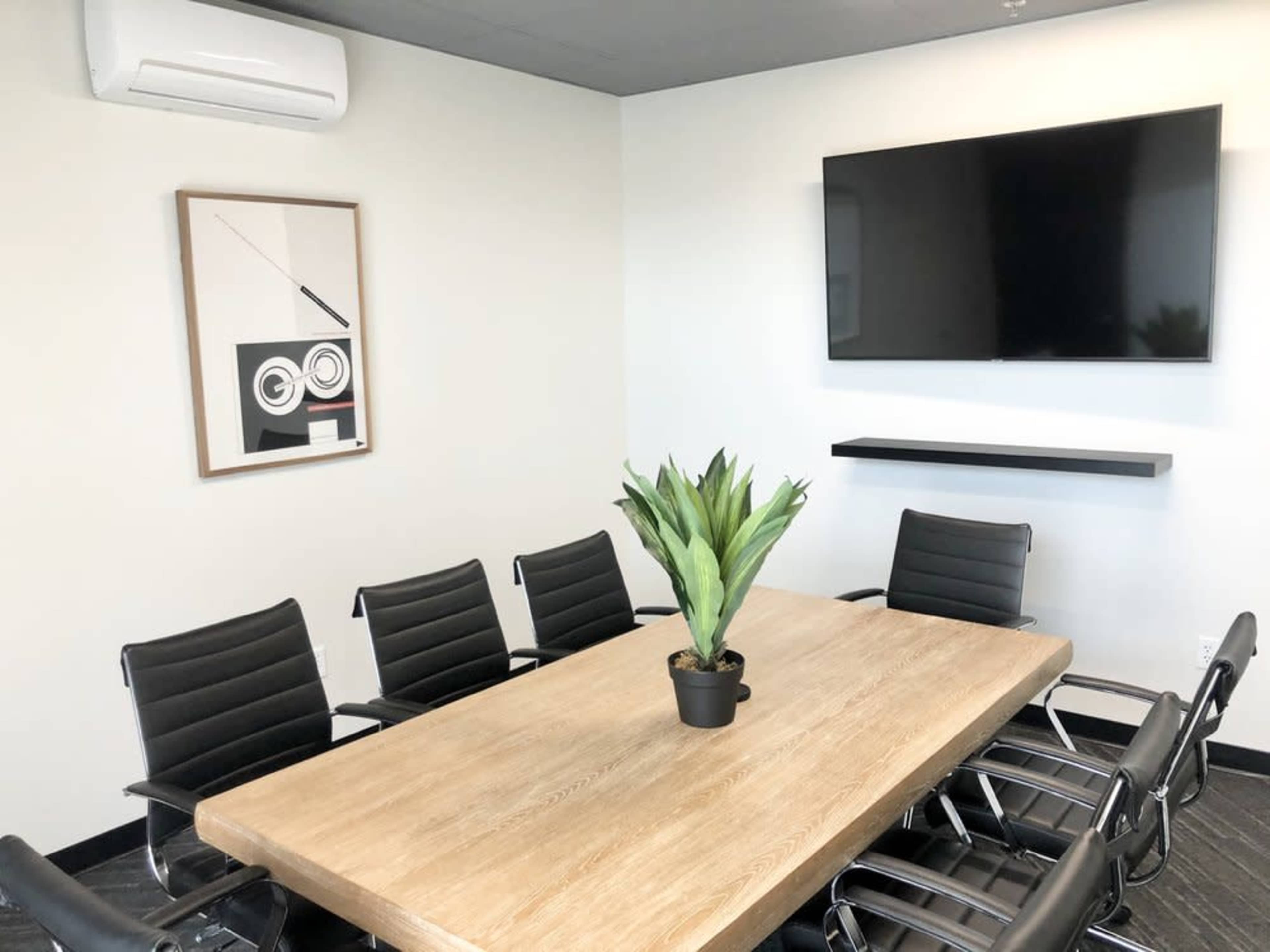 A modern conference room features a large wooden table surrounded by black chairs, with a television mounted on the wall and a plant in the center.