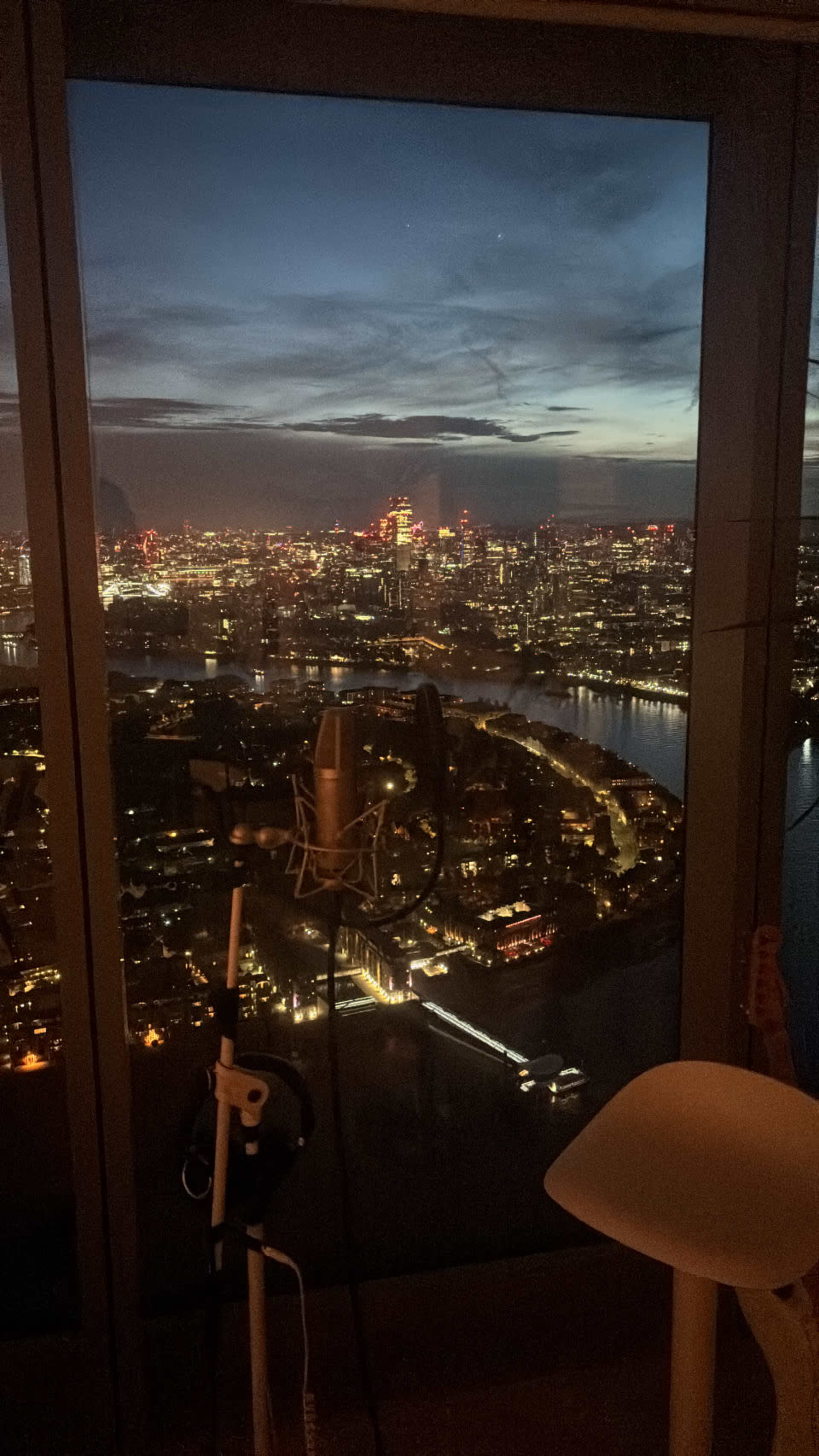 The image shows a recording microphone set up near a large window overlooking a city skyline at dusk, with vibrant lights reflecting on the water below.