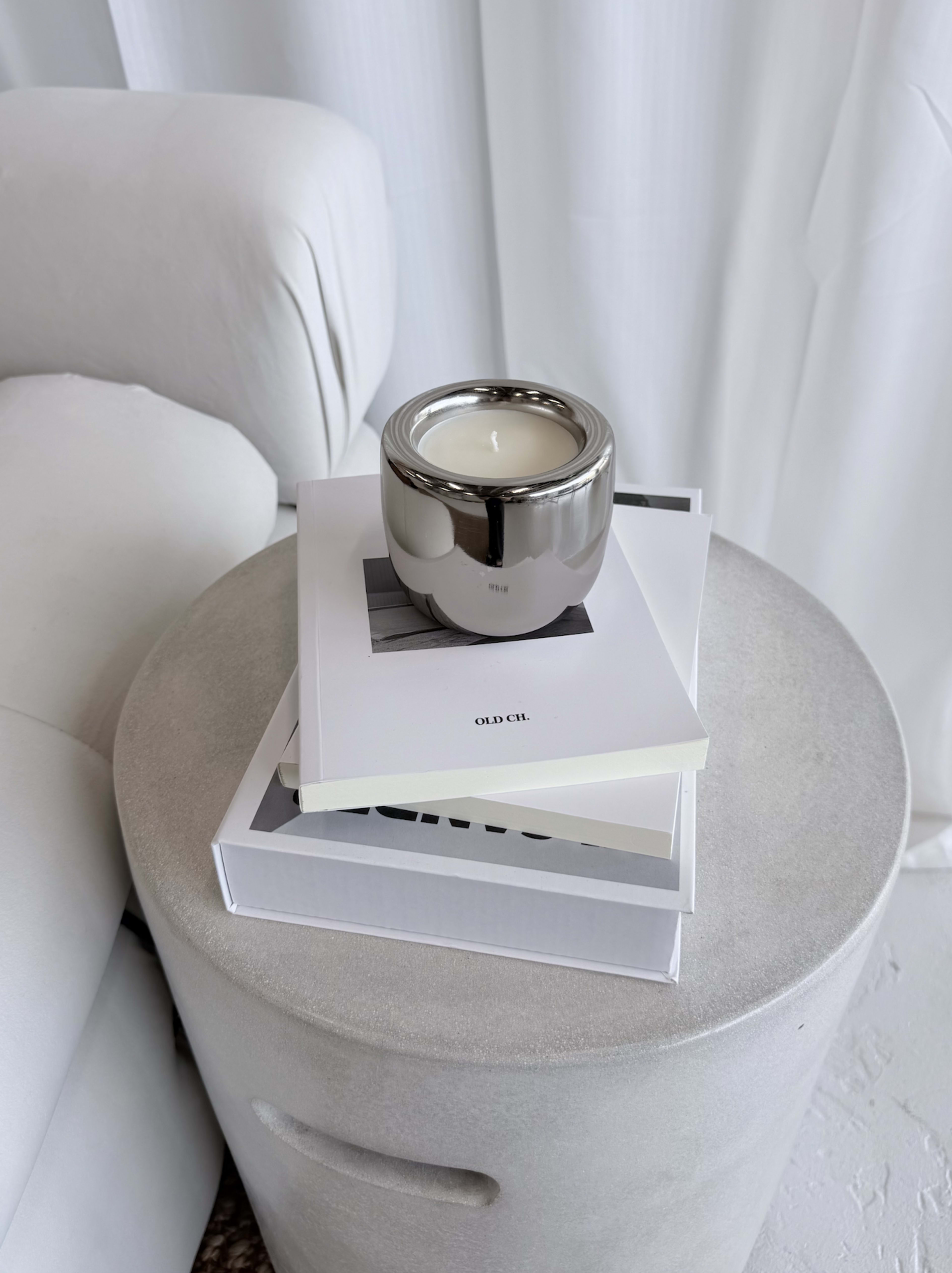 A silver candle sits on top of stacked books on a round concrete side table next to a light-colored sofa.
