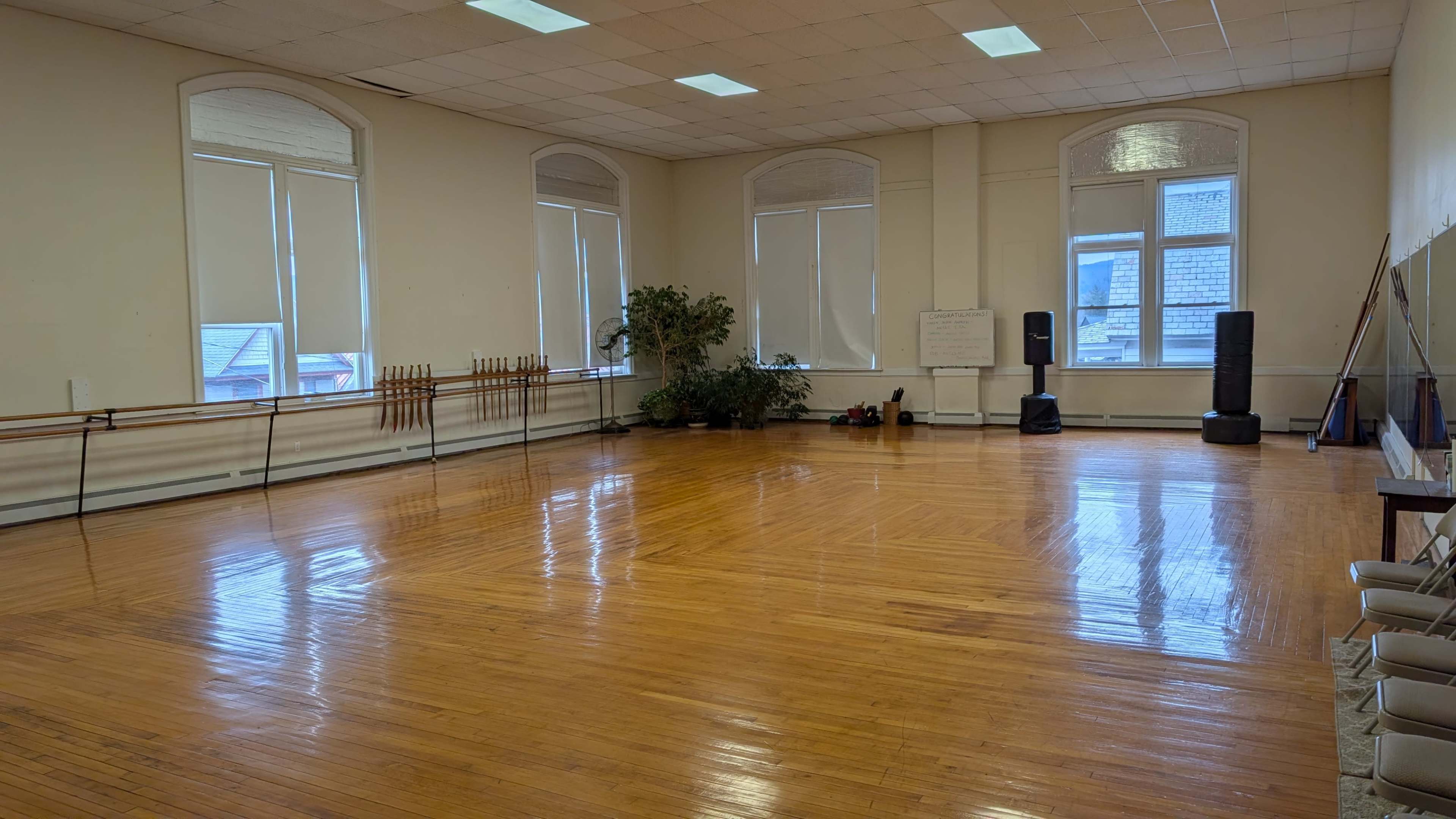 The image shows a spacious, empty dance studio with wooden flooring, large windows, and mirrors along the walls.