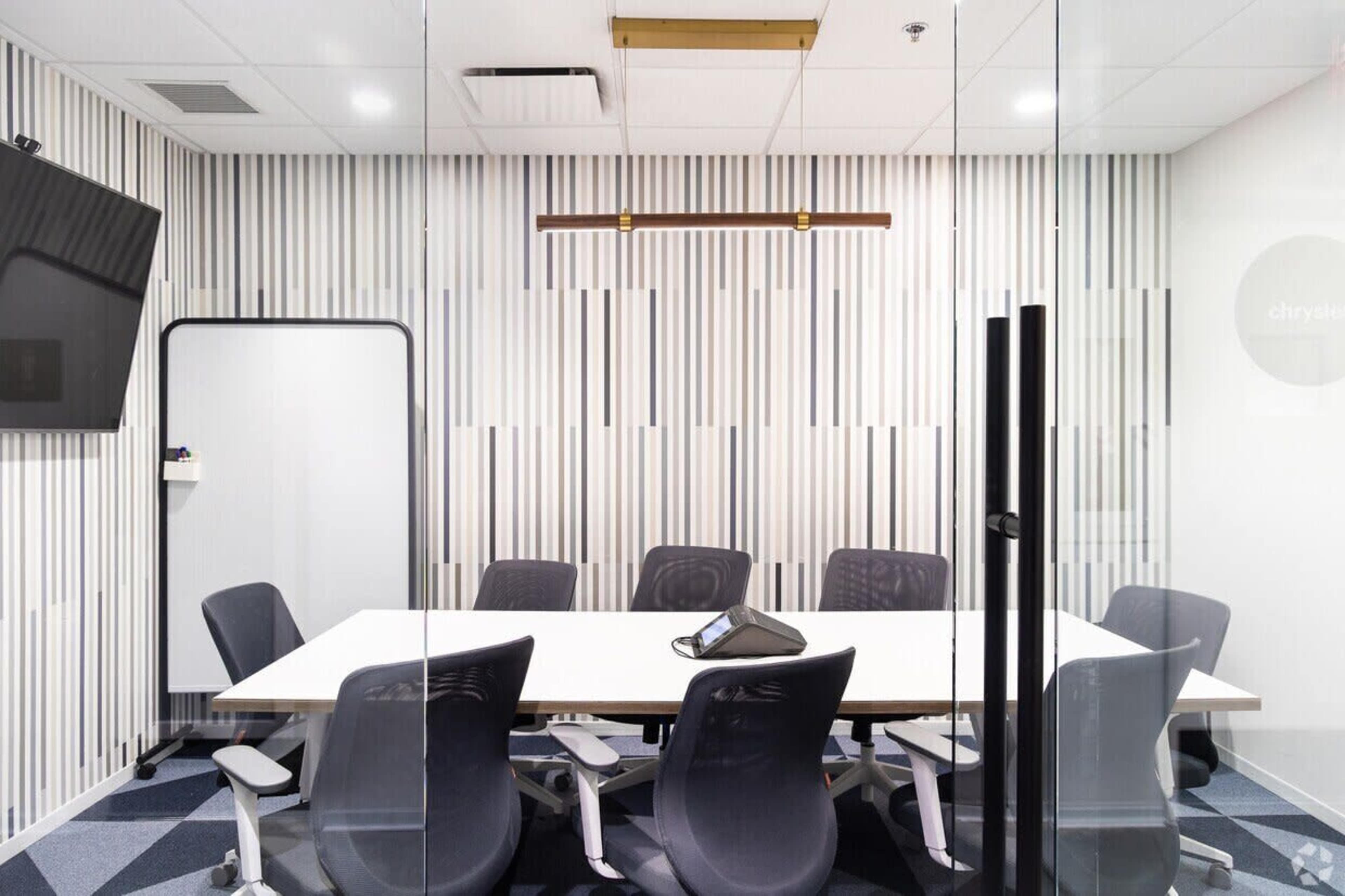 The image shows a modern conference room featuring a rectangular table surrounded by ergonomic chairs, with a wall-mounted screen and a glass partition.