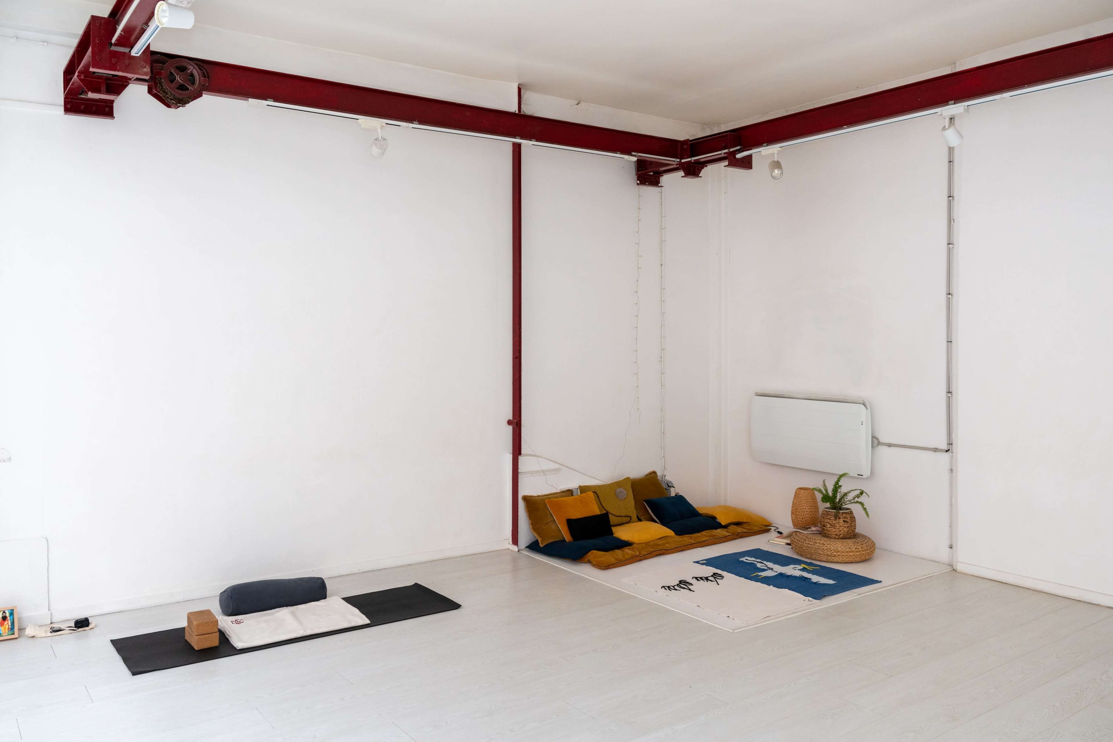 The image shows a minimalist interior space with a whitewashed wall, wooden floor, and two areas with folded blankets and cushions.