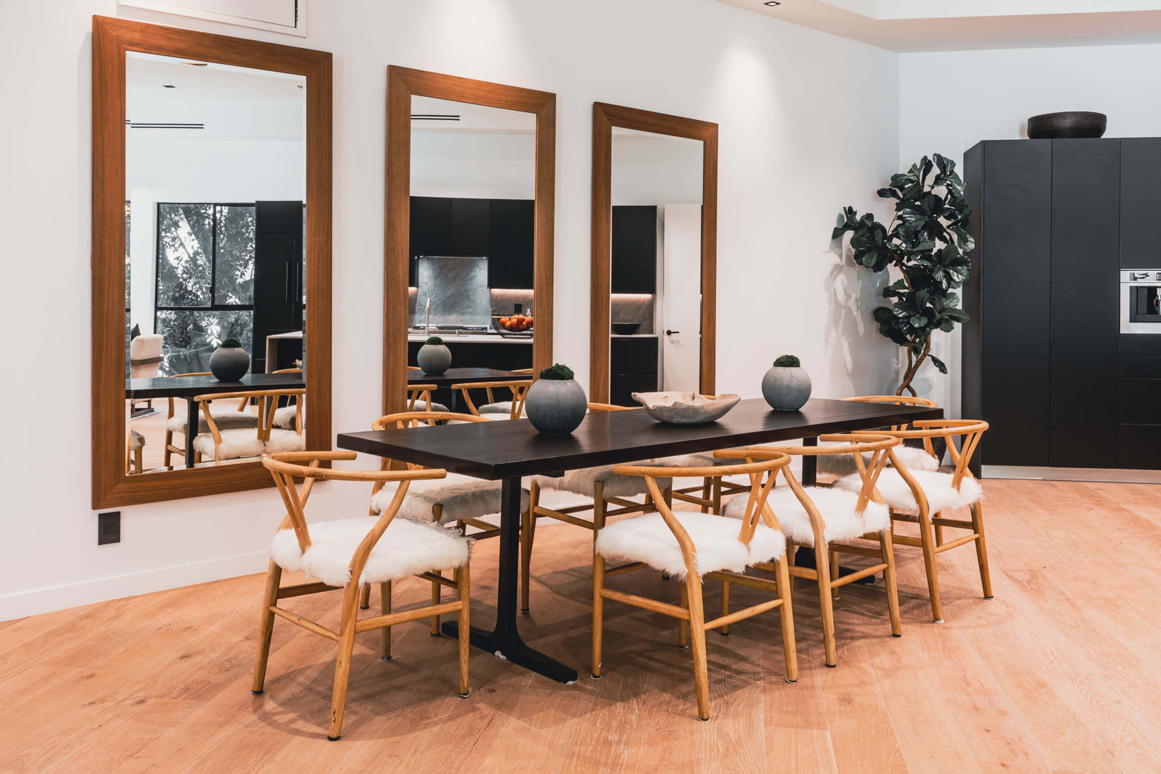 A modern dining area features a wooden table surrounded by light-colored chairs, with three large mirrors on the wall reflecting the space.