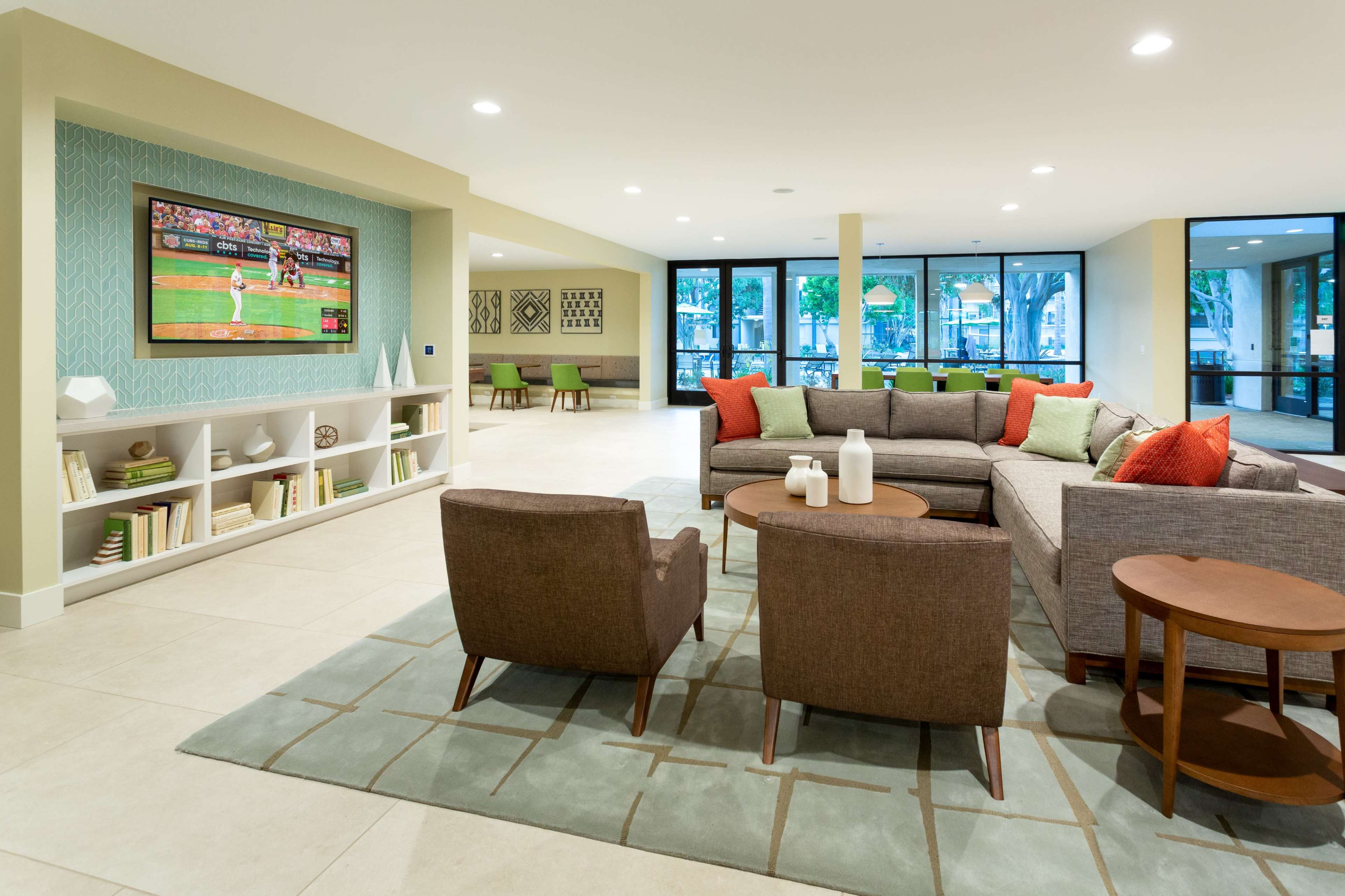 A modern living room with a large sectional sofa, two brown chairs, a coffee table, and a wall-mounted television displaying a baseball game, all set against a backdrop of large windows and neutral-colored walls.