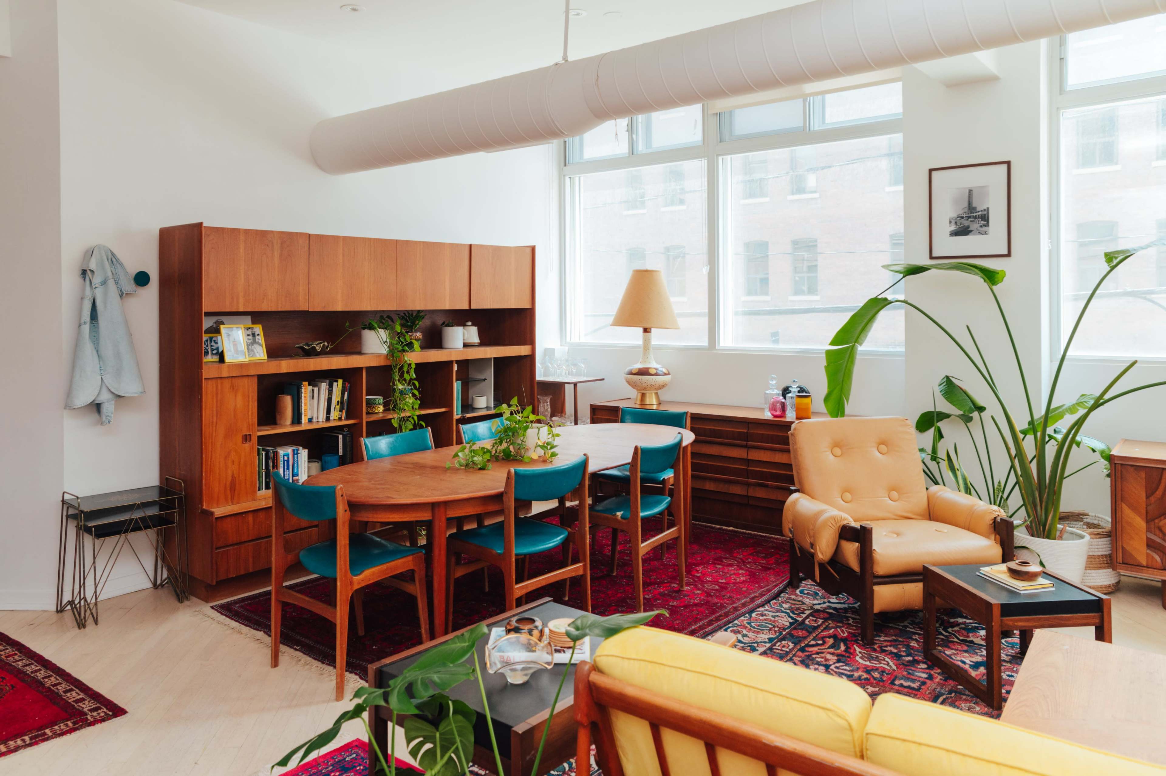 The image shows a bright, modern living room featuring a wooden bookshelf, a round dining table with teal chairs, and a retro armchair, all complemented by large windows and indoor plants.