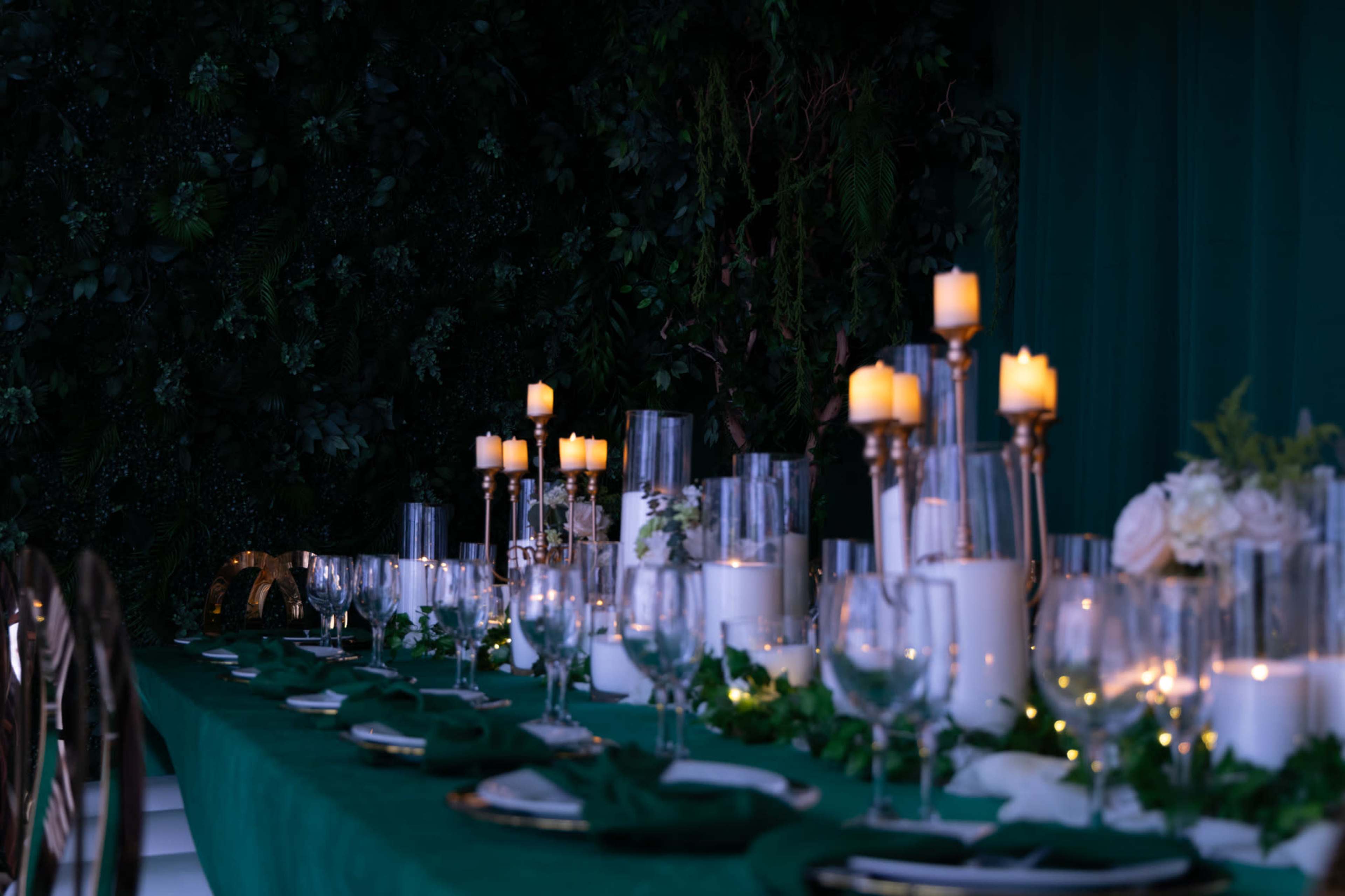 A long dining table is set with glassware, candles, and greenery against a dark green backdrop.