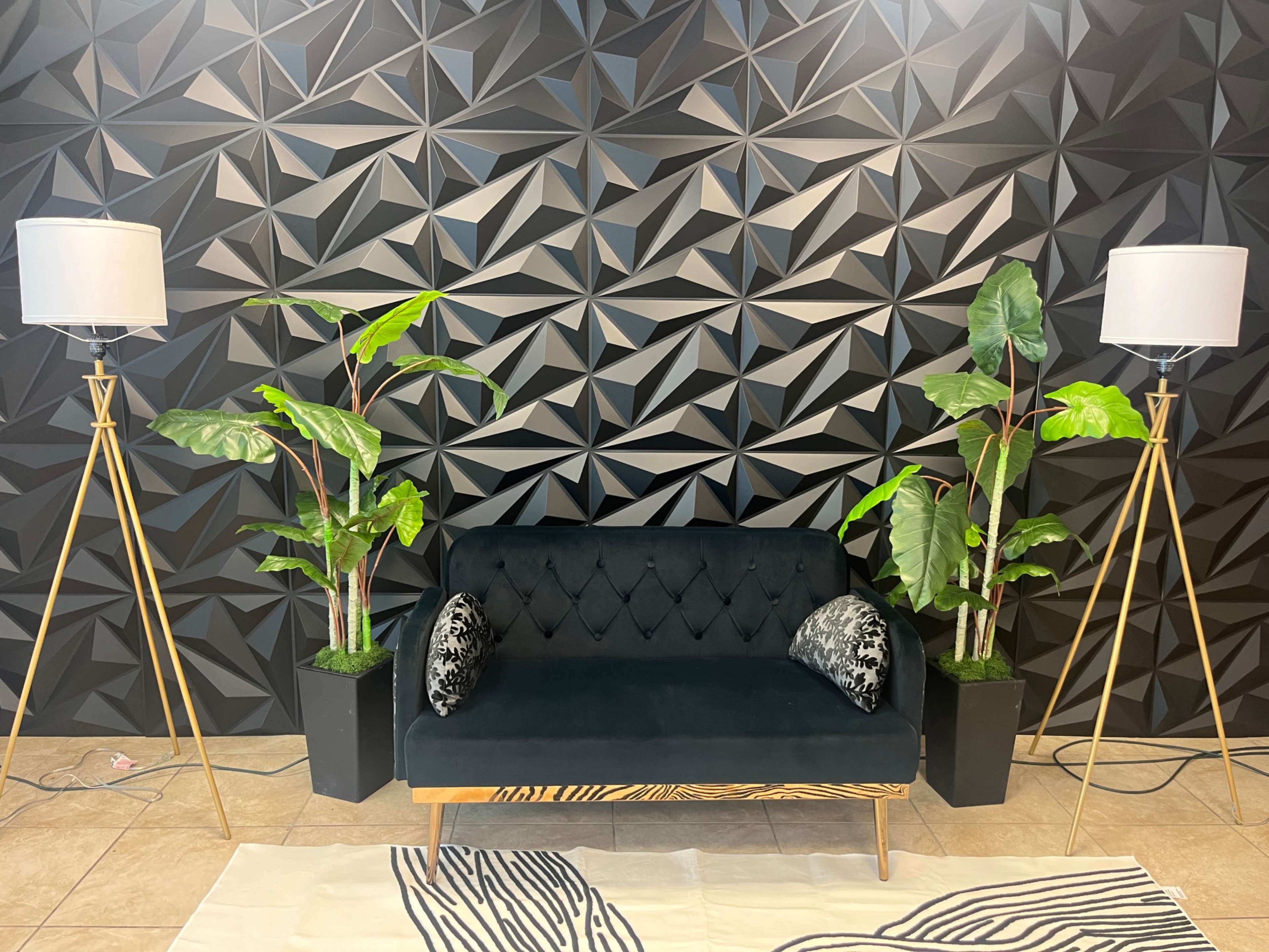 A black sofa with decorative pillows is positioned against a textured black geometric wall, flanked by two tall floor lamps and potted plants.