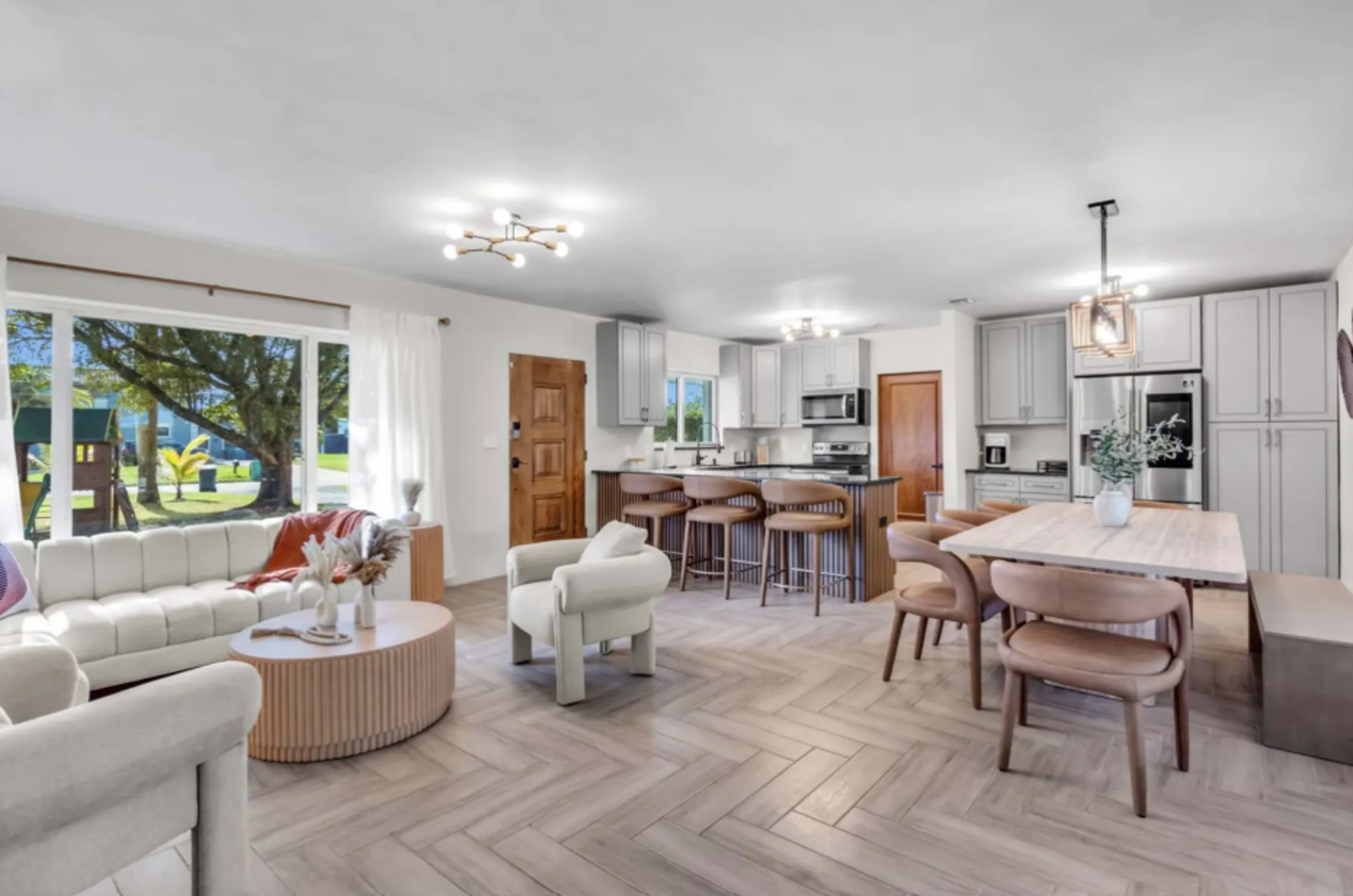 The image shows a modern living space with an open kitchen, dining area, and seating arrangement featuring light-colored furniture and wooden accents.
