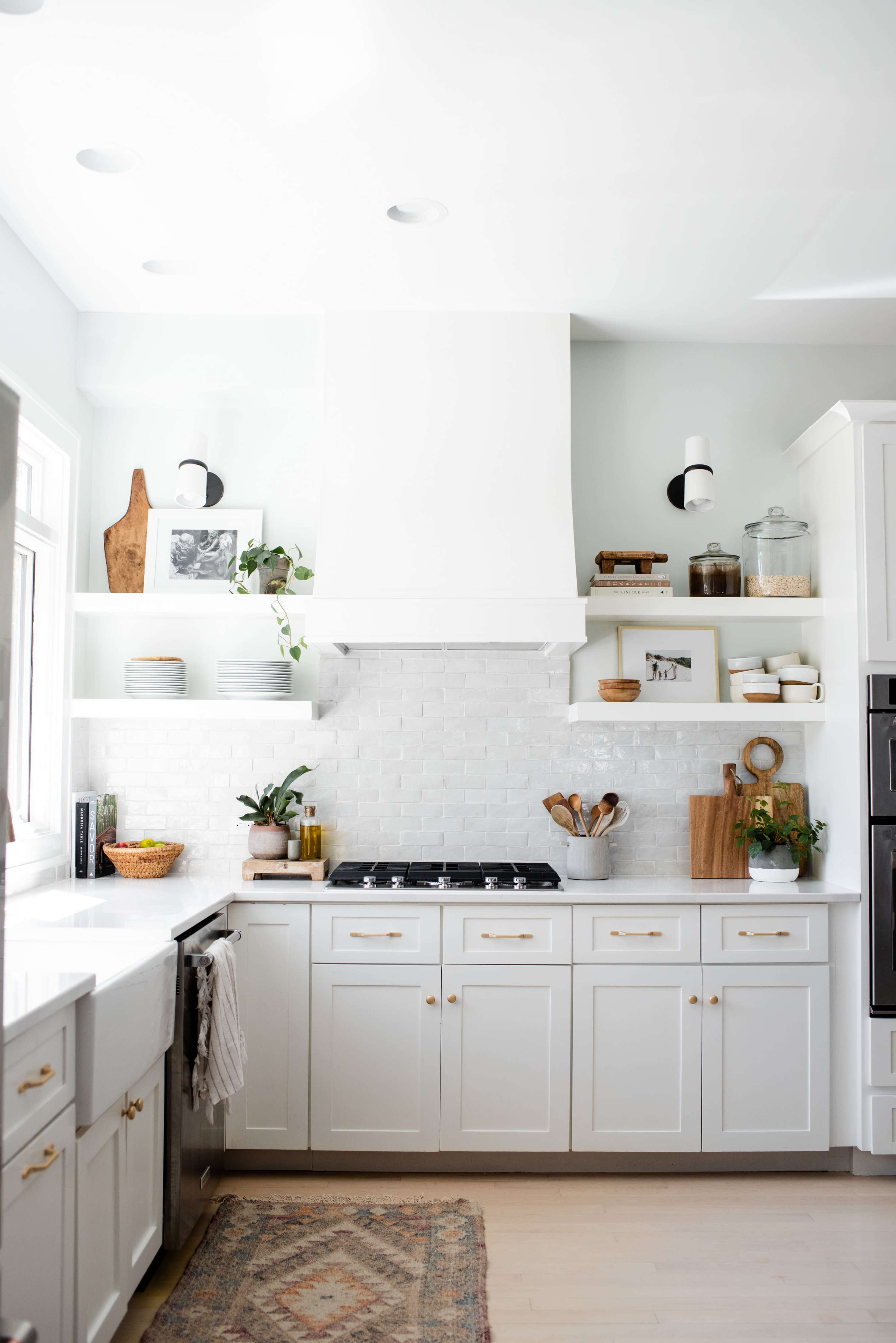 The image shows a modern kitchen with white cabinetry, a large hood above the stove, open shelves displaying plants and kitchen items, and a patterned rug on the floor.