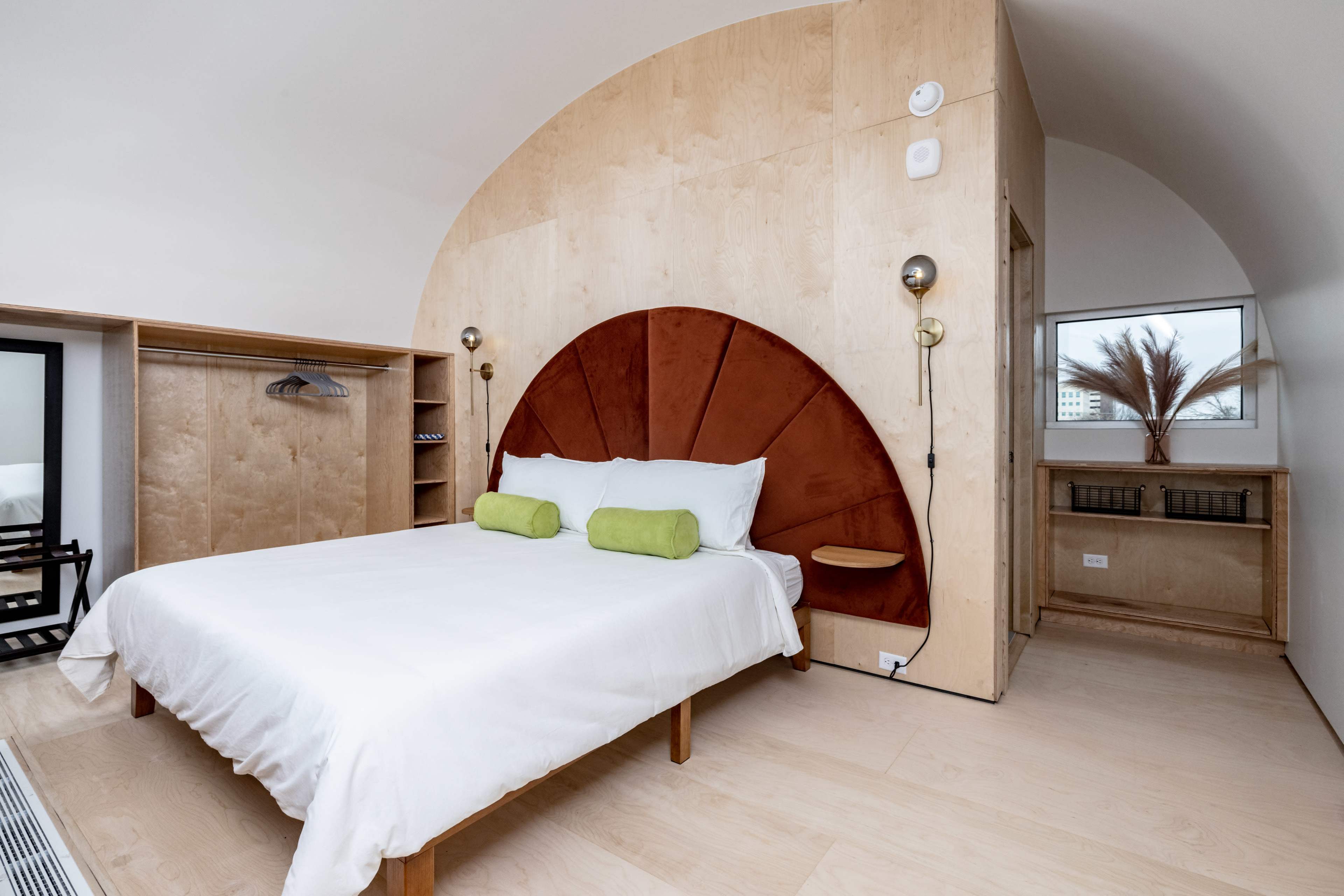 A minimalist bedroom with a wooden bed frame, a unique arched headboard, and simple decor, featuring a window and shelving unit.