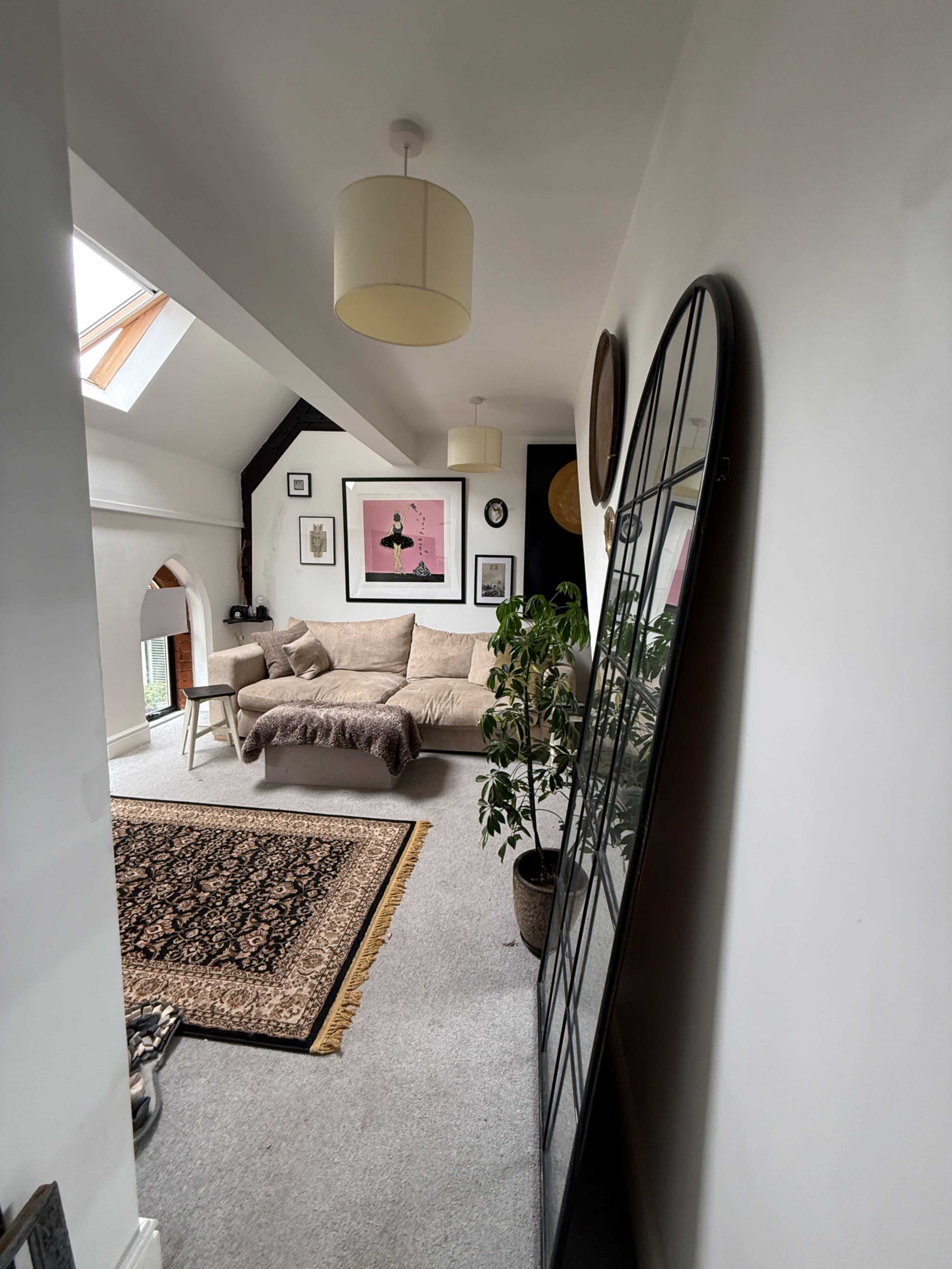 The image shows a spacious living room featuring a large mirror, a beige couch, a decorative rug, and various wall art pieces.