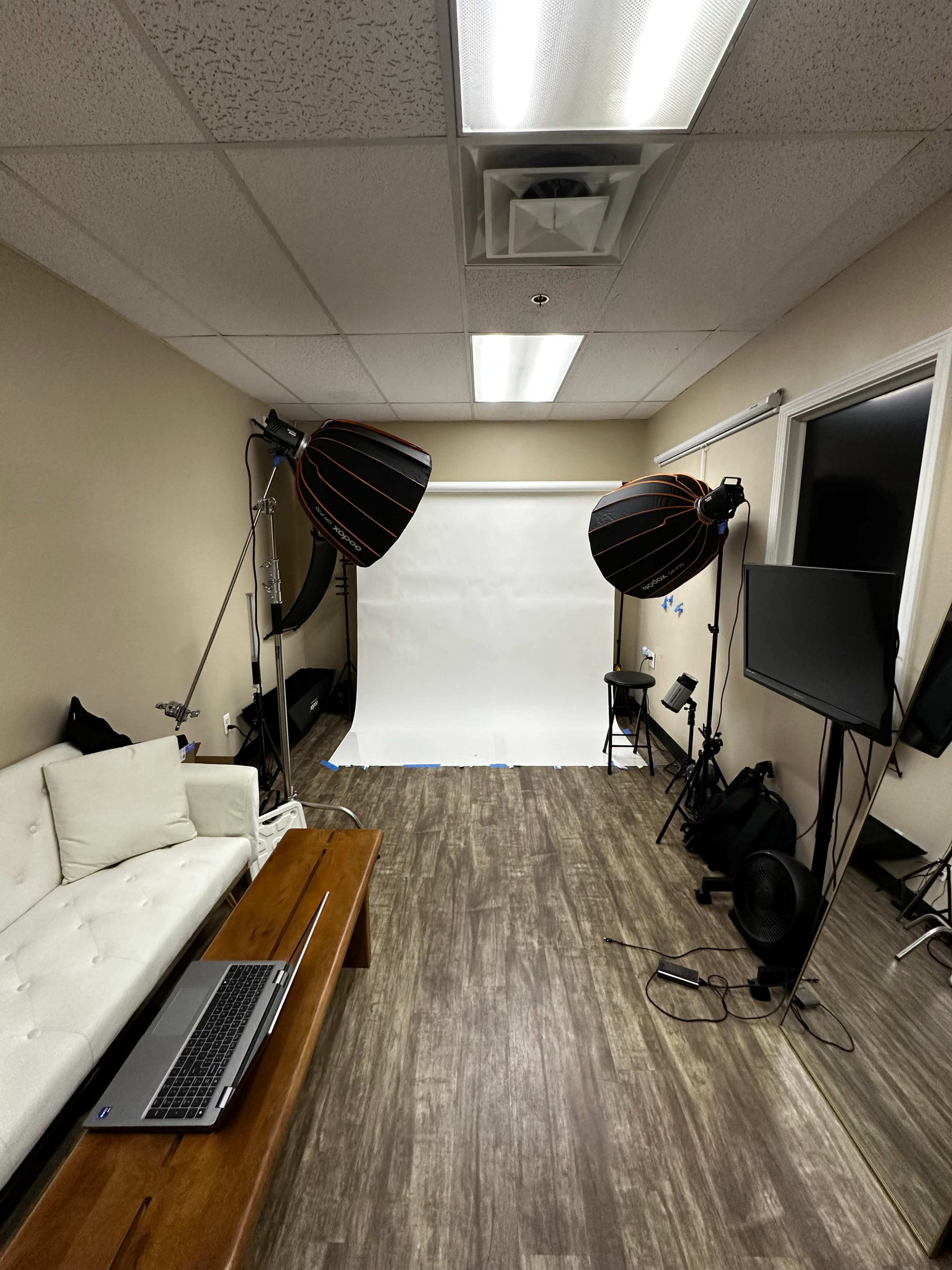 A photography studio with a white backdrop, two large softbox lights, a laptop on a table, and a wall-mounted screen.