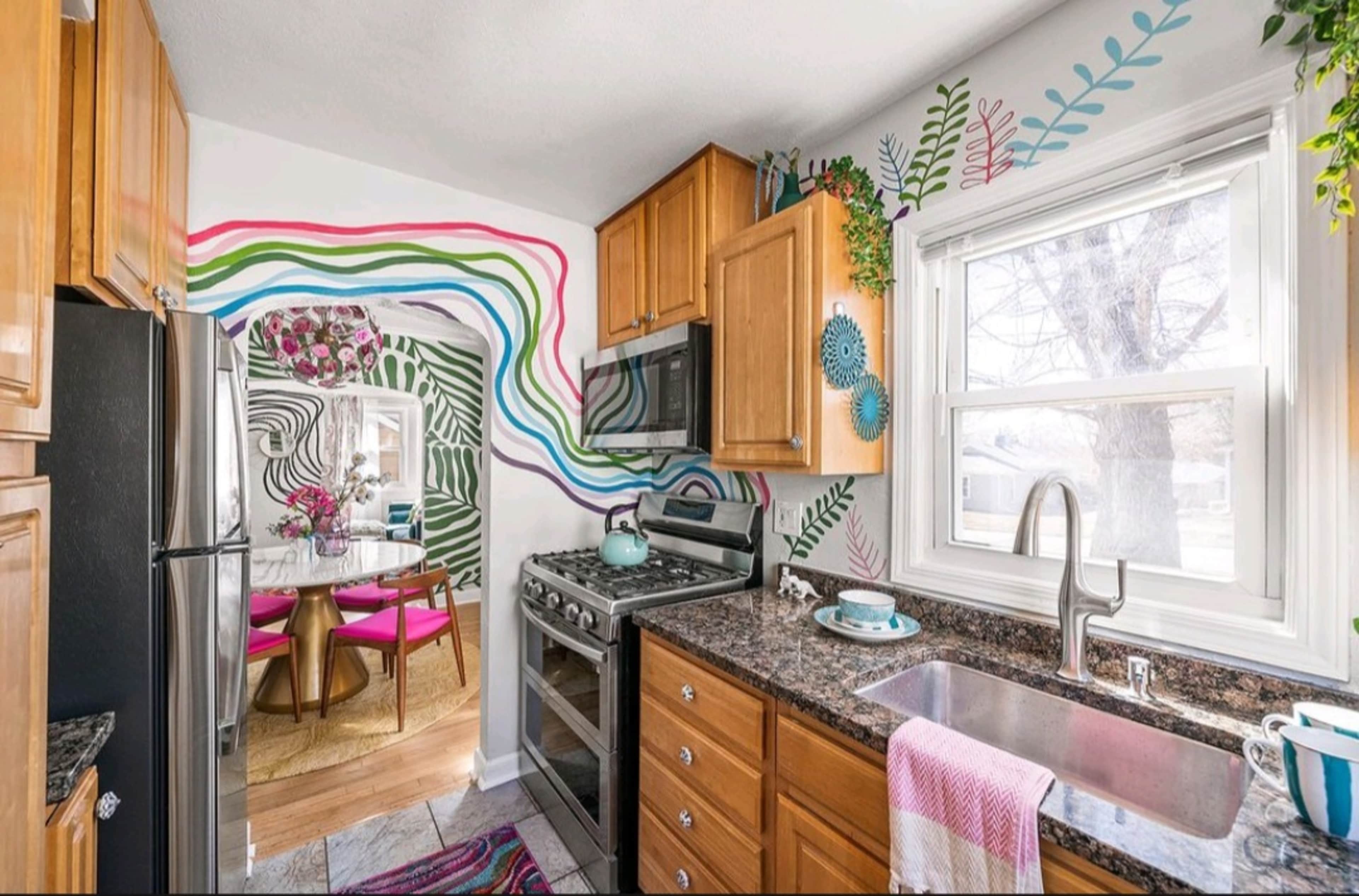 A colorful kitchen features wooden cabinets, a stainless steel stove, and a sink, with vibrant wall art and a view into a dining area with a round table and pink chairs.