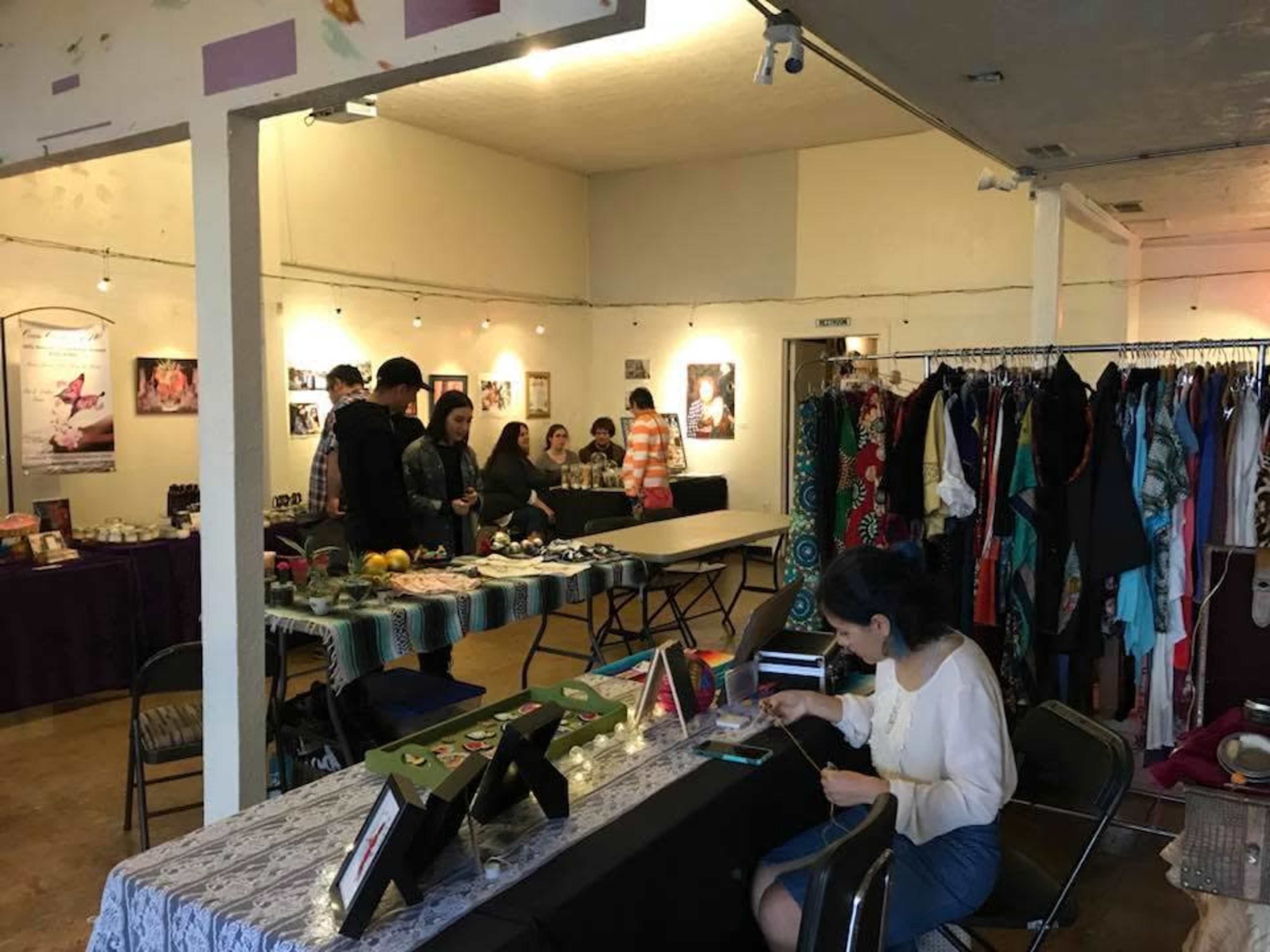 The image shows a community event space with tables displaying various items, people interacting, and racks of clothing along the walls.