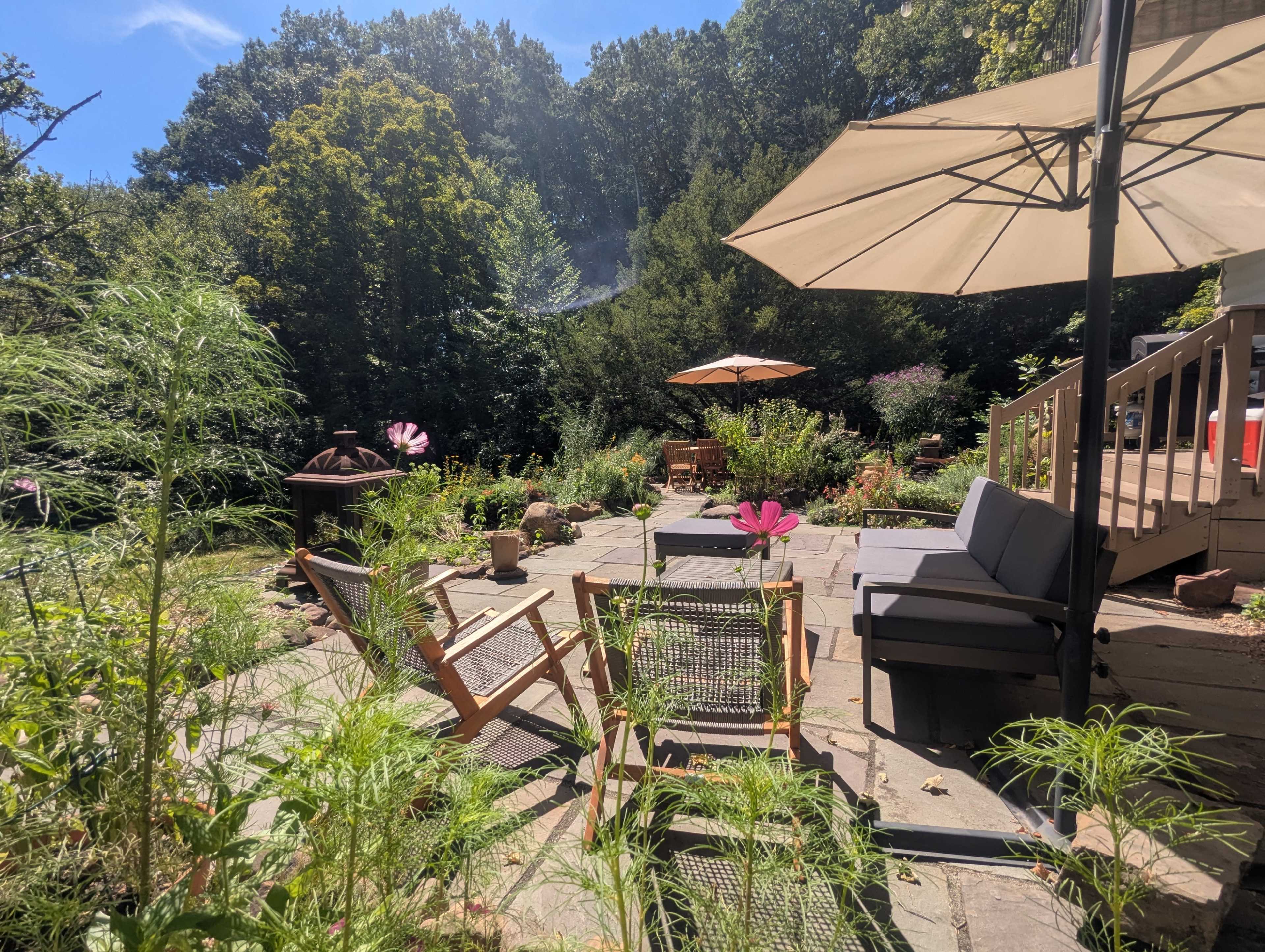 The image shows a patio area with outdoor seating, surrounded by greenery and a large umbrella for shade.