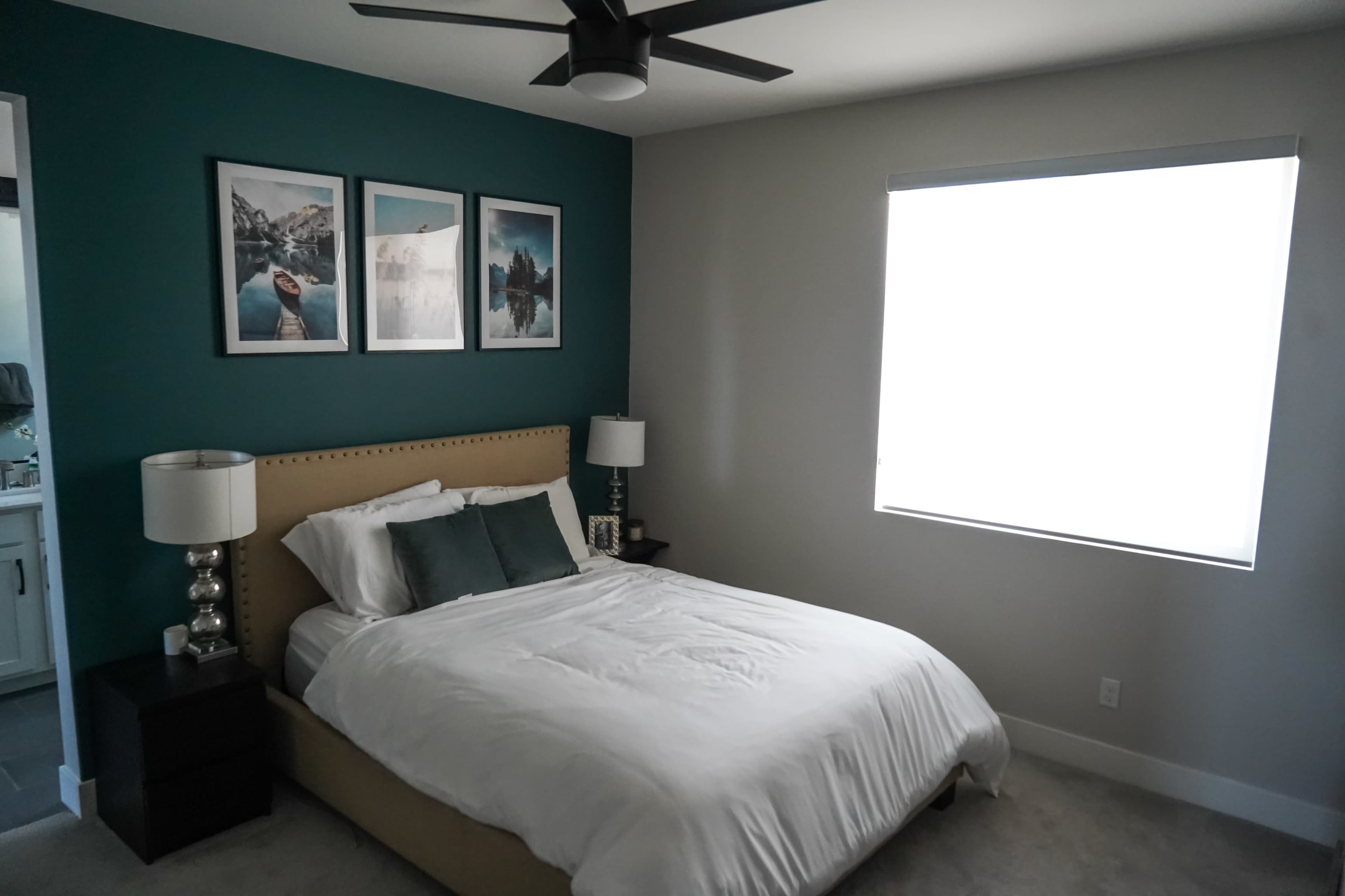A neatly arranged bedroom features a bed with white linens, two bedside lamps, a window with sheer coverings, and framed landscape photos on the teal wall.