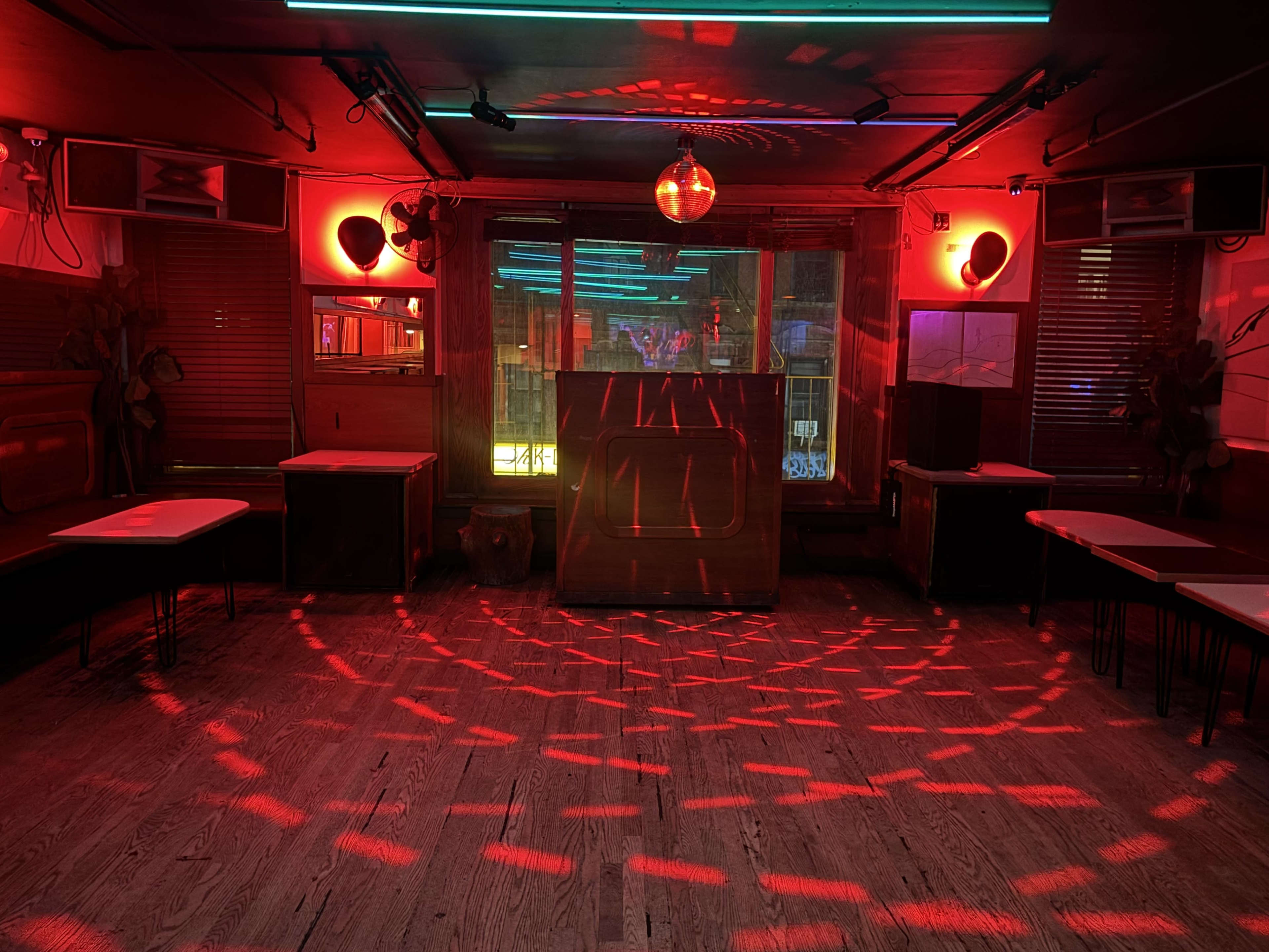 The image shows a dimly lit dance floor with red lighting, a disco ball, and a bar area surrounded by tables and chairs.