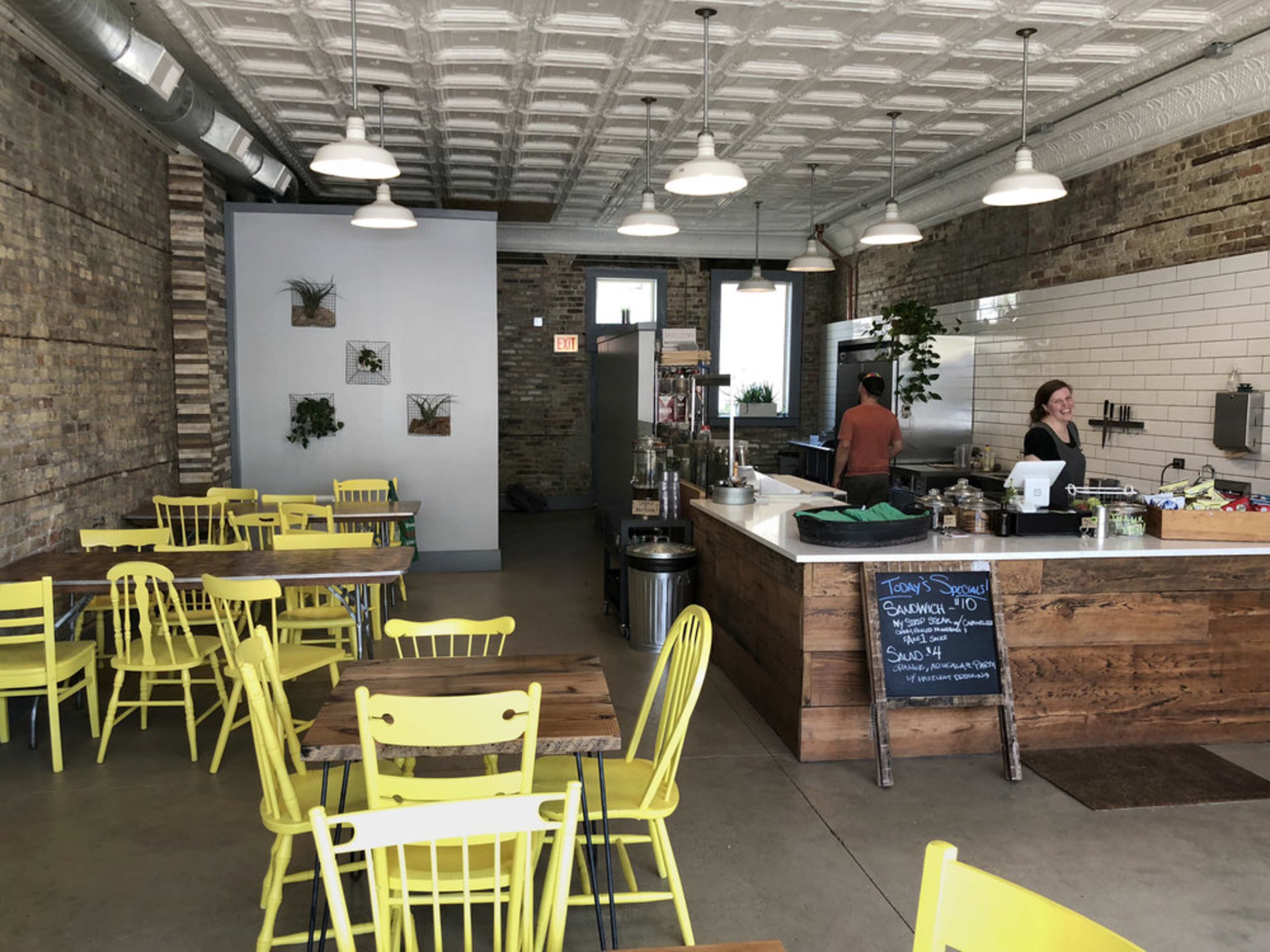 A cafe interior with yellow chairs and wooden tables, featuring a service counter in the background and exposed brick walls.
