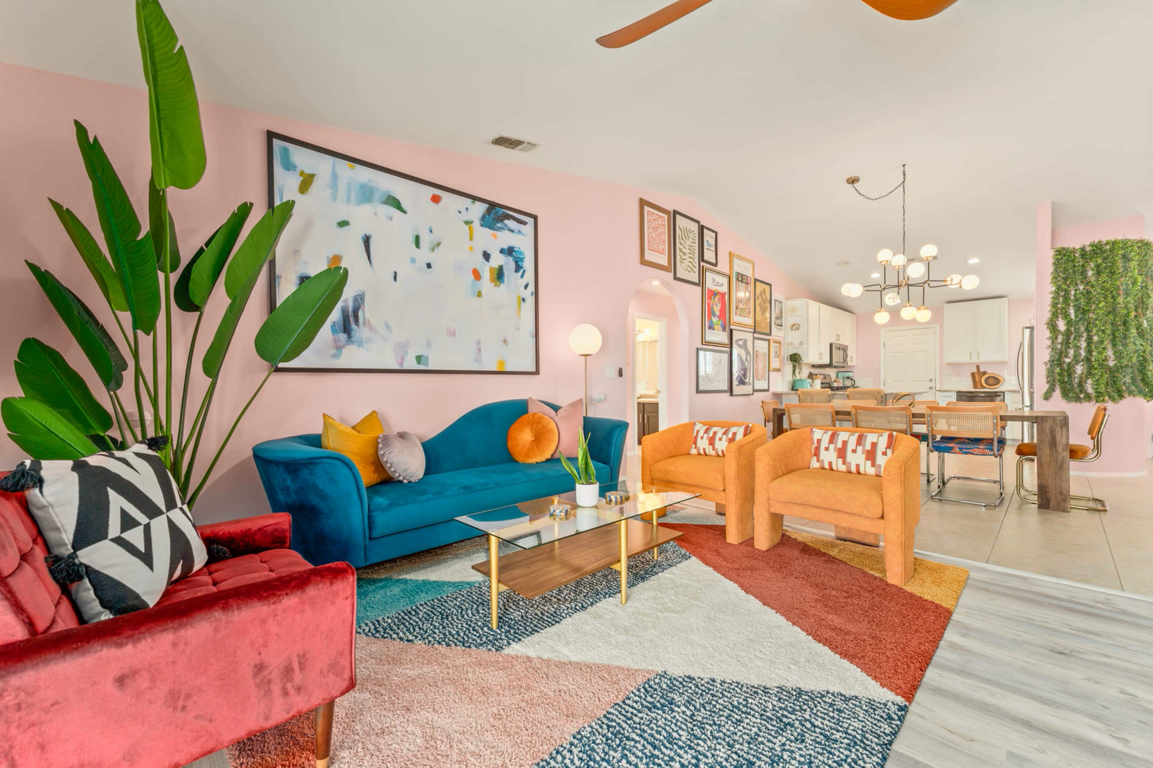 The scene features a colorful living room with a blue sofa, a red chair, and orange armchairs, adorned with a large abstract painting and various framed artworks on the walls.