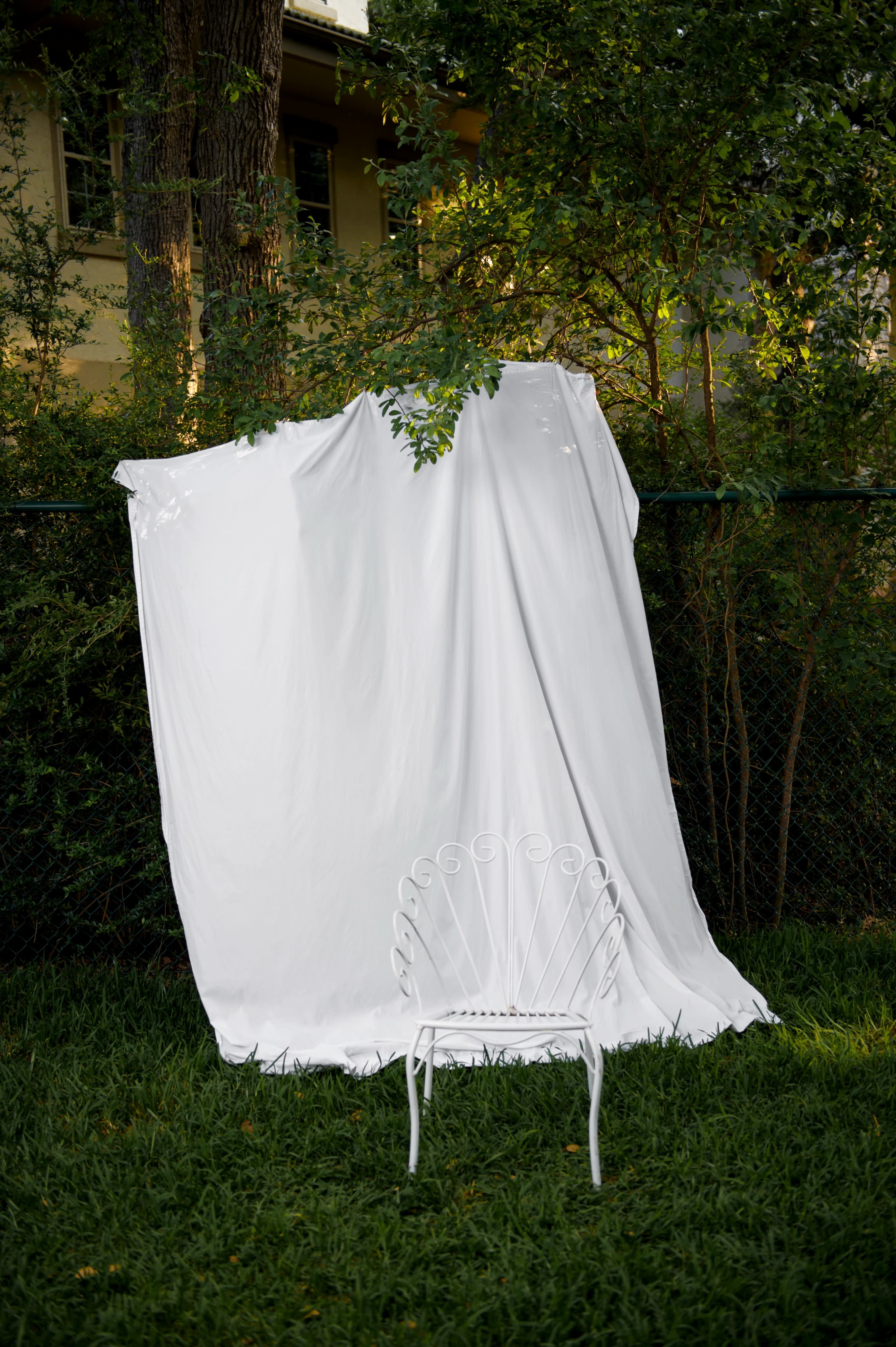 A white sheet is draped over a fence beside a white metal chair in a grassy area.