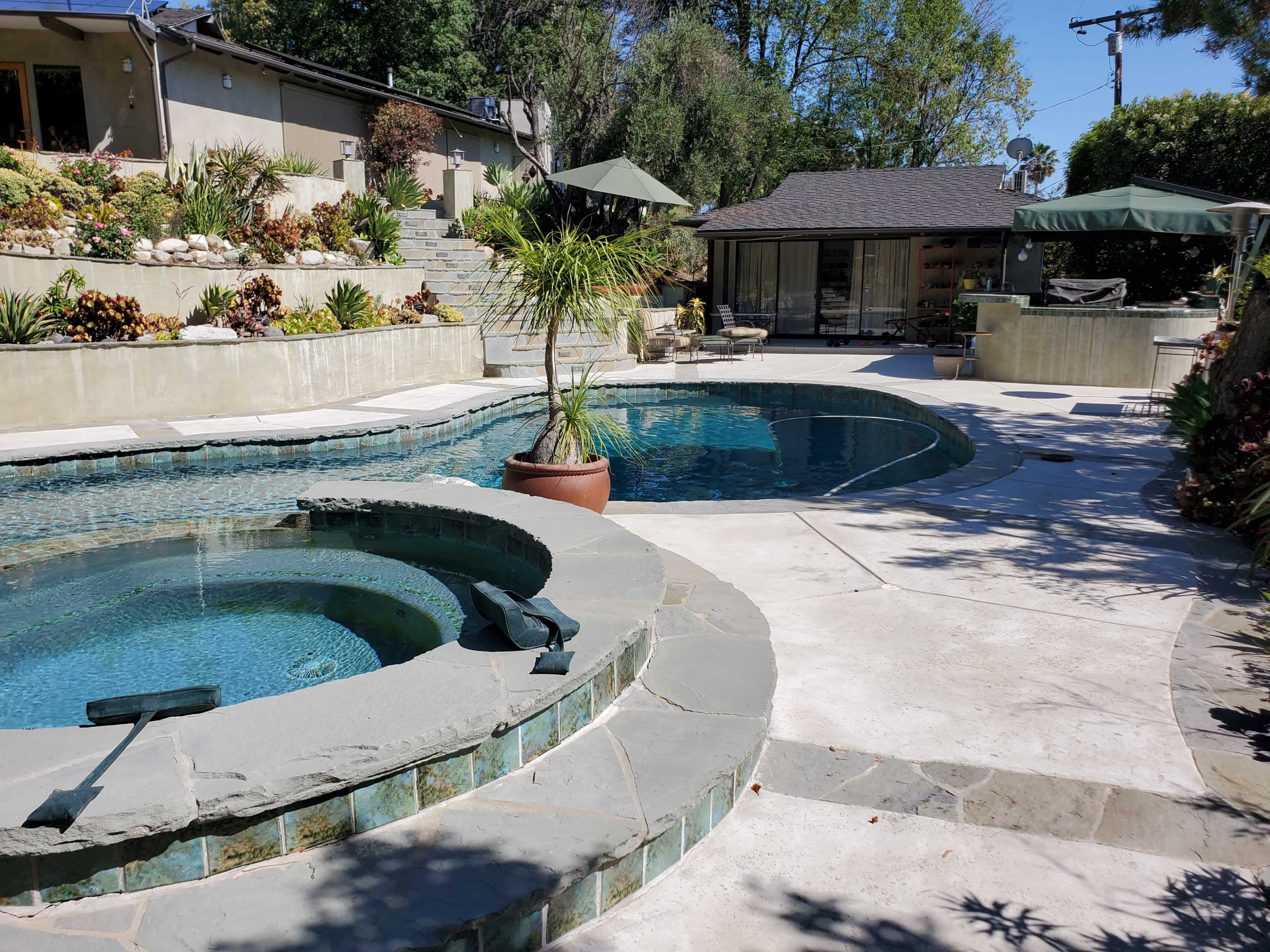 A landscaped pool area featuring a hot tub, a larger swimming pool, and surrounding greenery with a patio.