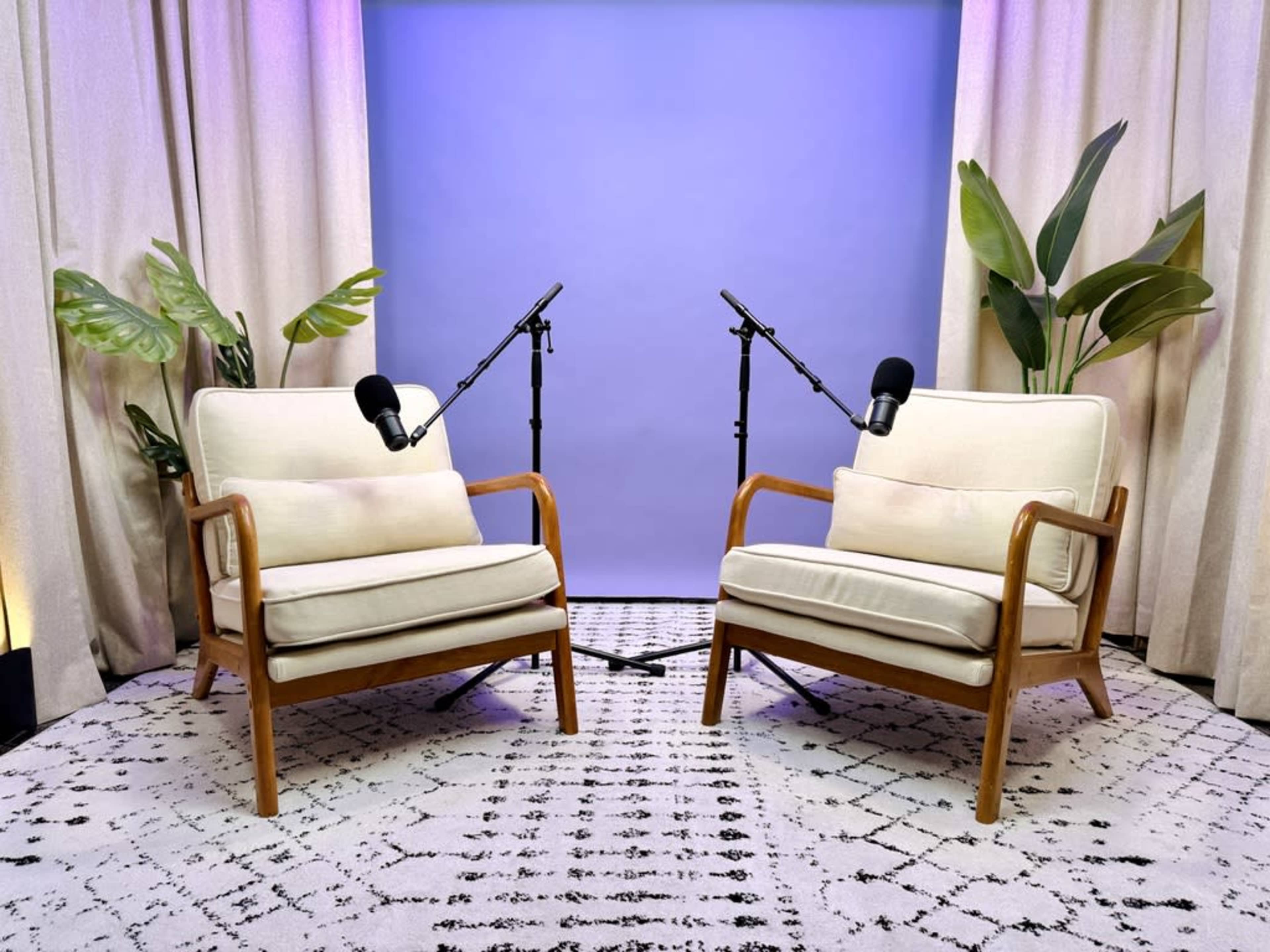 The image shows two cream-colored armchairs facing each other, with microphones on stands positioned between them, set against a light blue backdrop and surrounded by curtain drapes and potted plants.