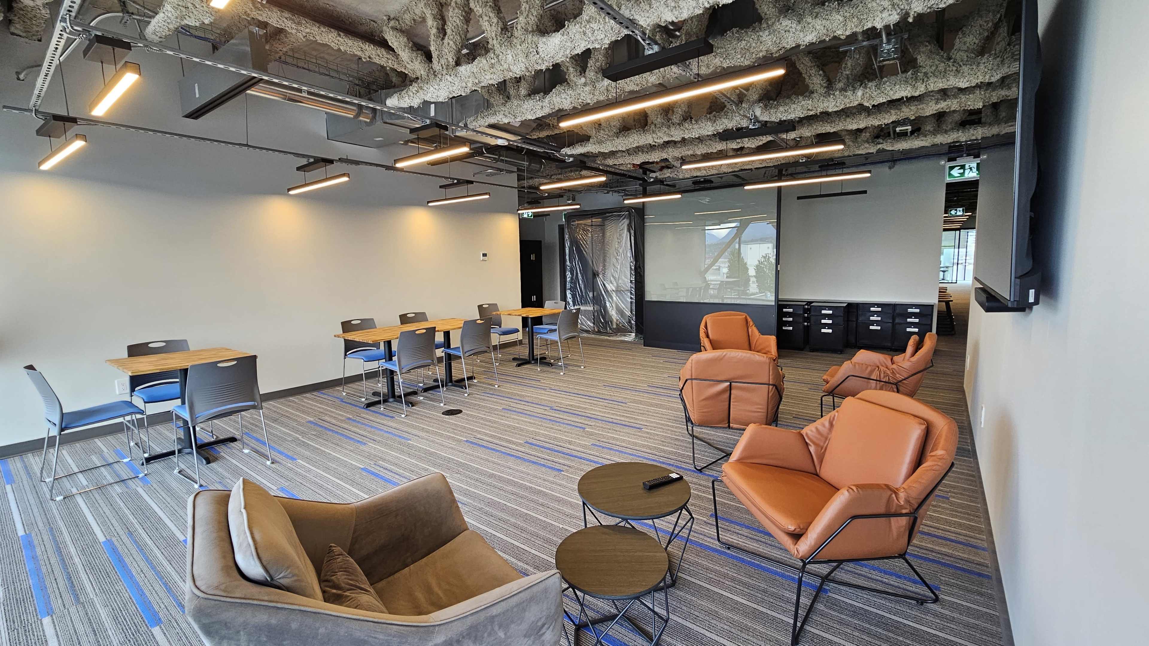 The image shows a modern office space featuring several tables and chairs, two lounge chairs, and a television mounted on the wall.