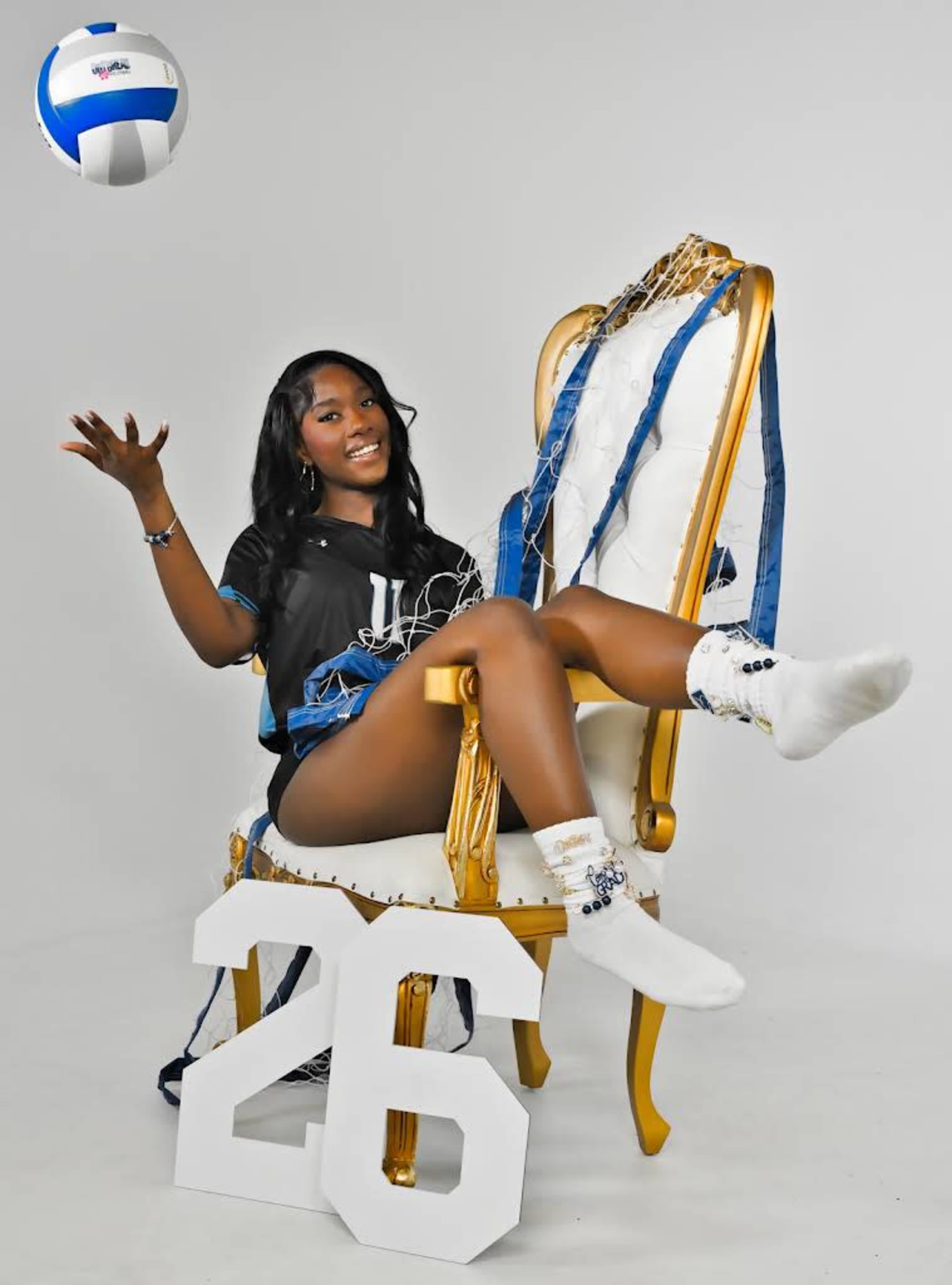 A person in athletic attire sits on a decorative chair, playfully tossing a volleyball while surrounded by a net and a large number "26."