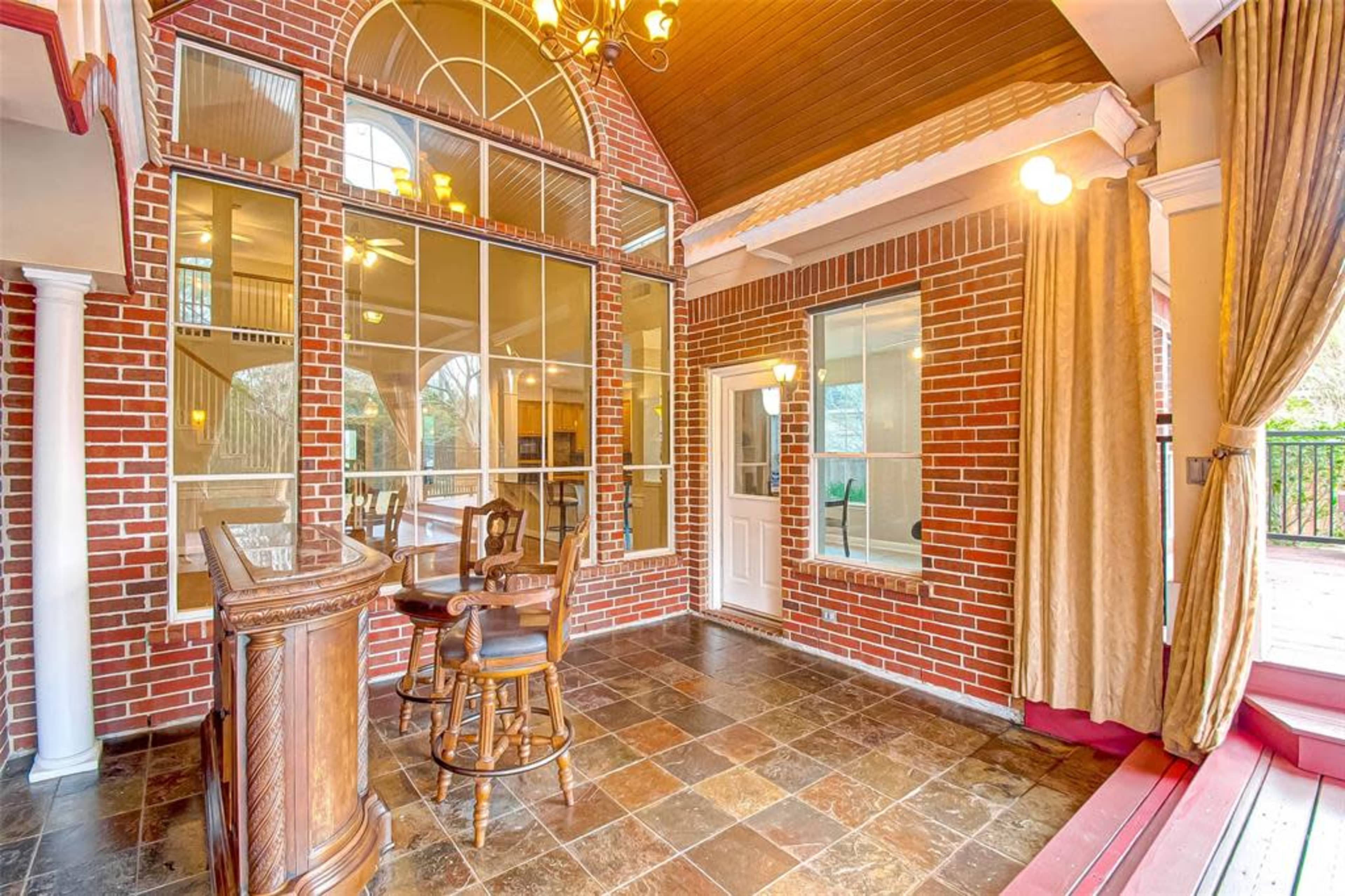 A spacious room with brick walls, a bar area, large windows, and a door leading outside, accented by a high ceiling and decorative lighting.