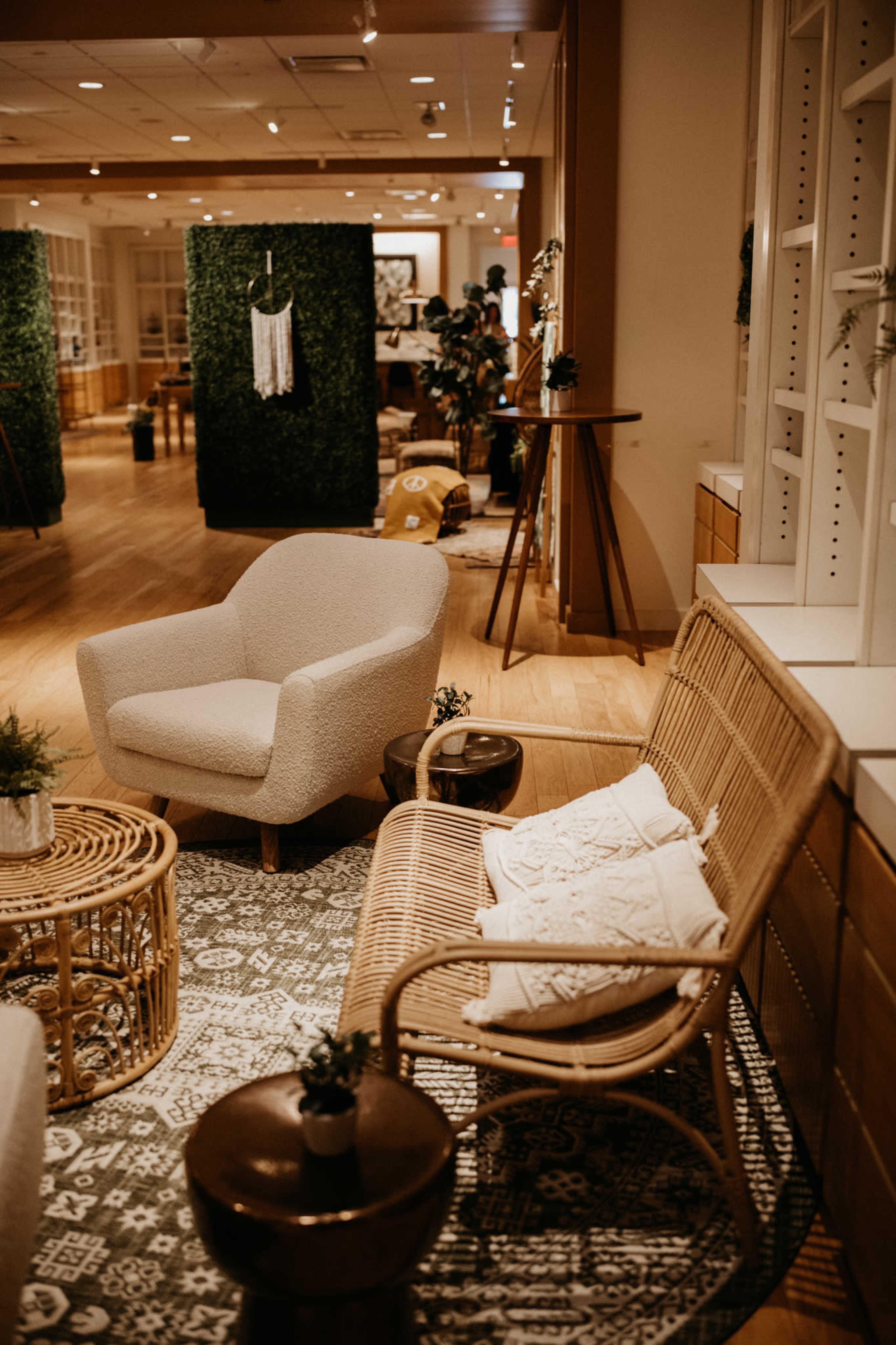 A cozy interior space featuring a light-colored armchair, a rattan chair, a round wooden table, and decorative plants against a backdrop of green foliage panels.