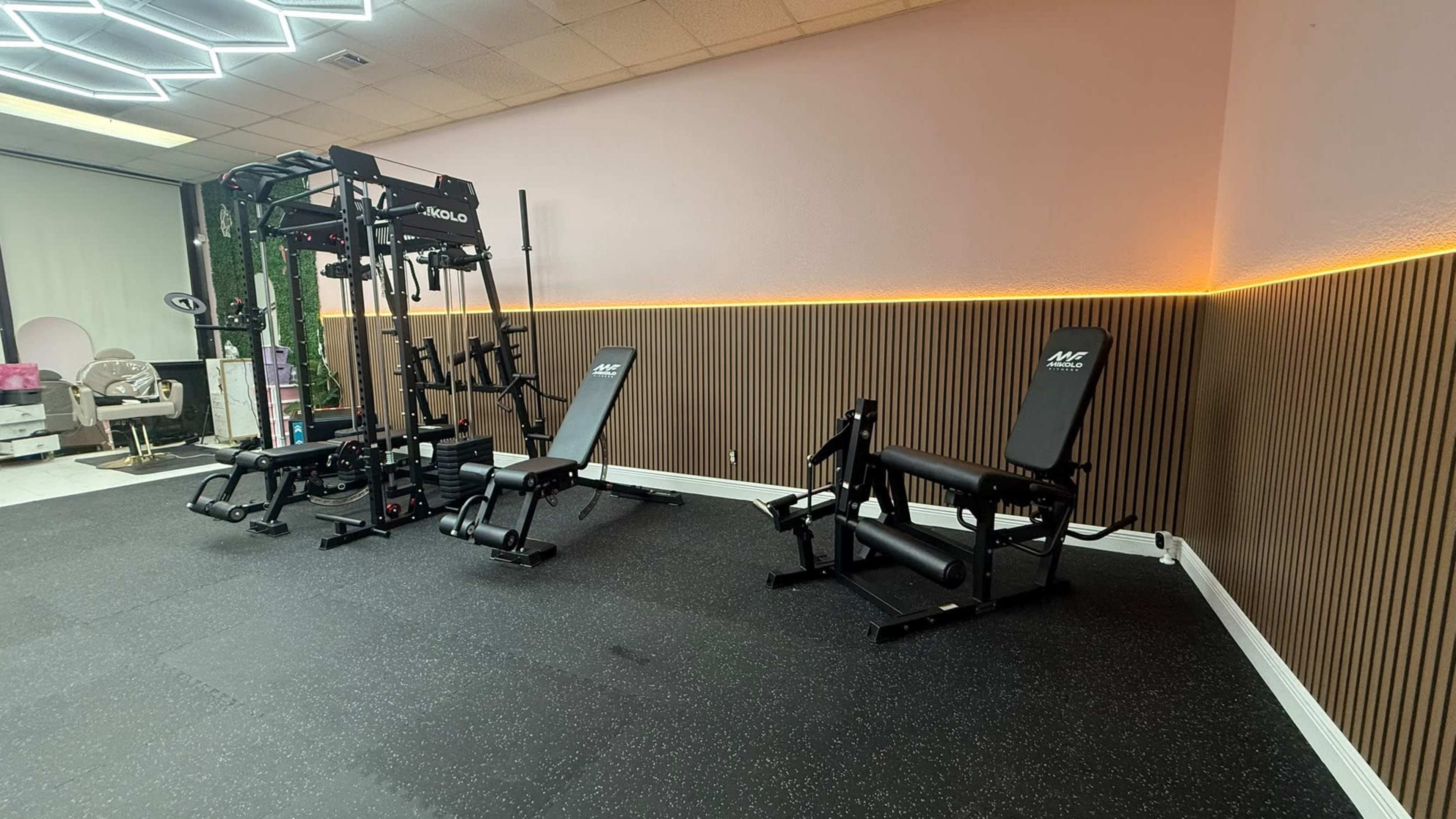 The image shows a gym area featuring a weight training machine on the left and two adjustable benches on the right, all set against a light pink wall with ambient lighting.