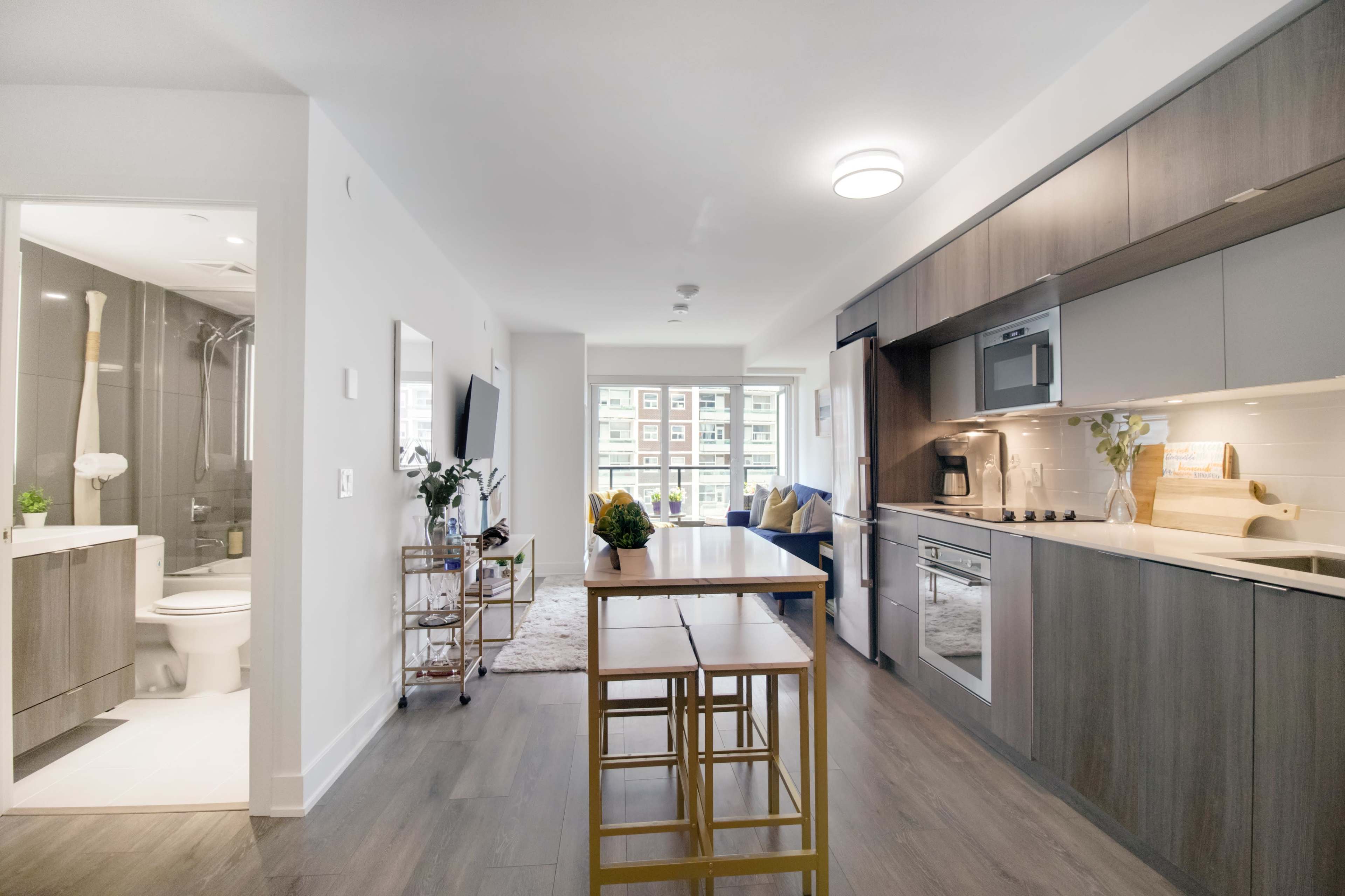 A modern, open-concept living space featuring a kitchen with stainless steel appliances, a dining table, and a bathroom visible to the left.