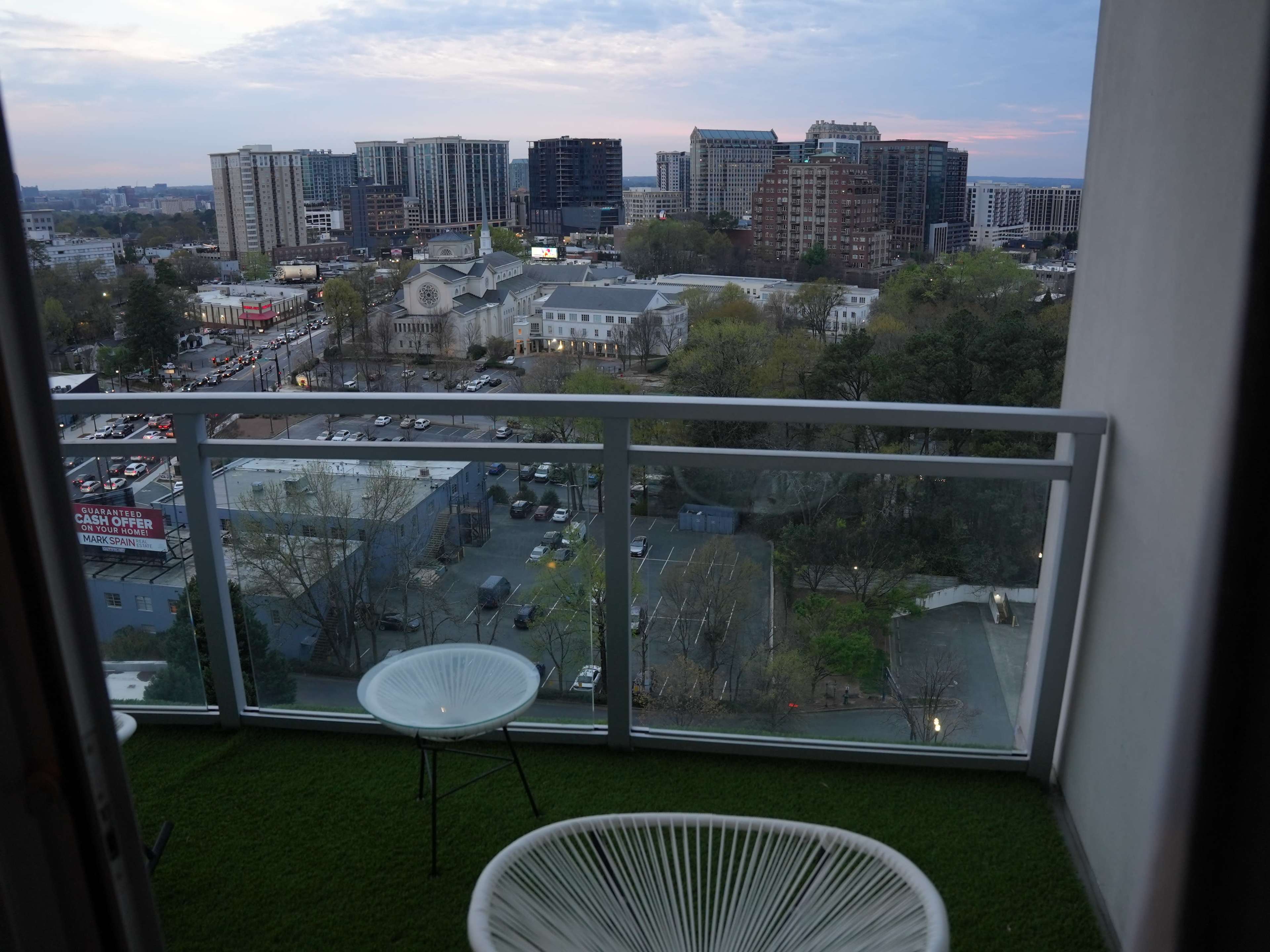 A balcony with white chairs overlooks a city skyline and parking lot at dusk.