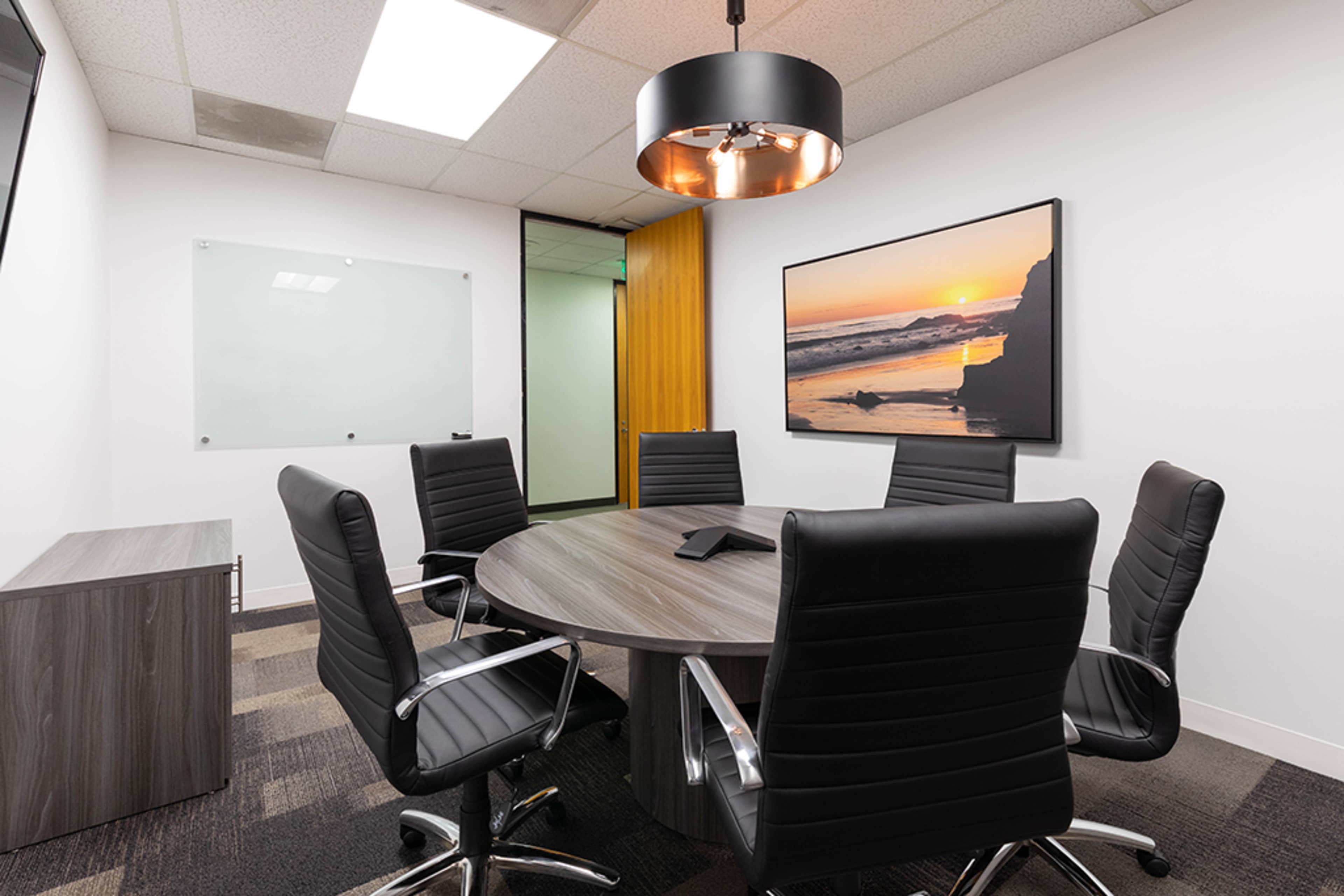 A conference room features a circular table surrounded by black leather chairs, with a large wall-mounted picture of a sunset and a glass whiteboard nearby.