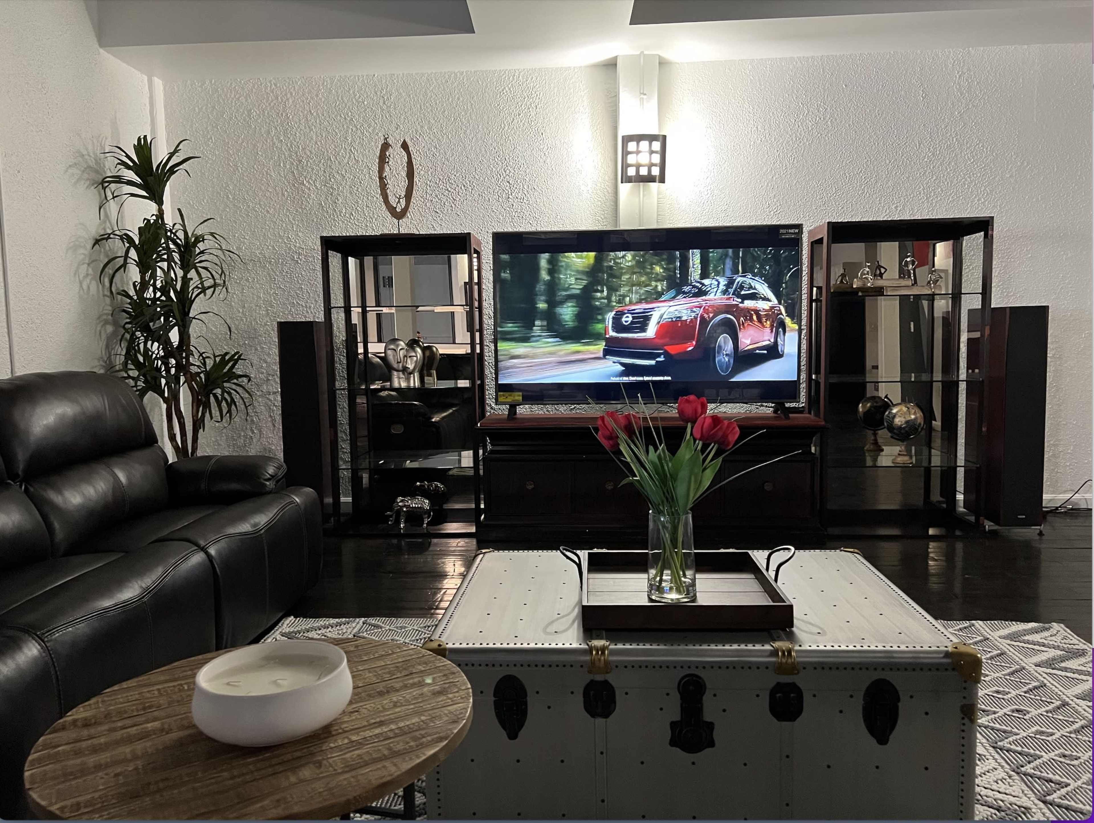 A modern living room features a black leather sofa, a wooden coffee table, a decorative trunk, and a large television displaying a car commercial, surrounded by glass display cabinets and a vase of red tulips.