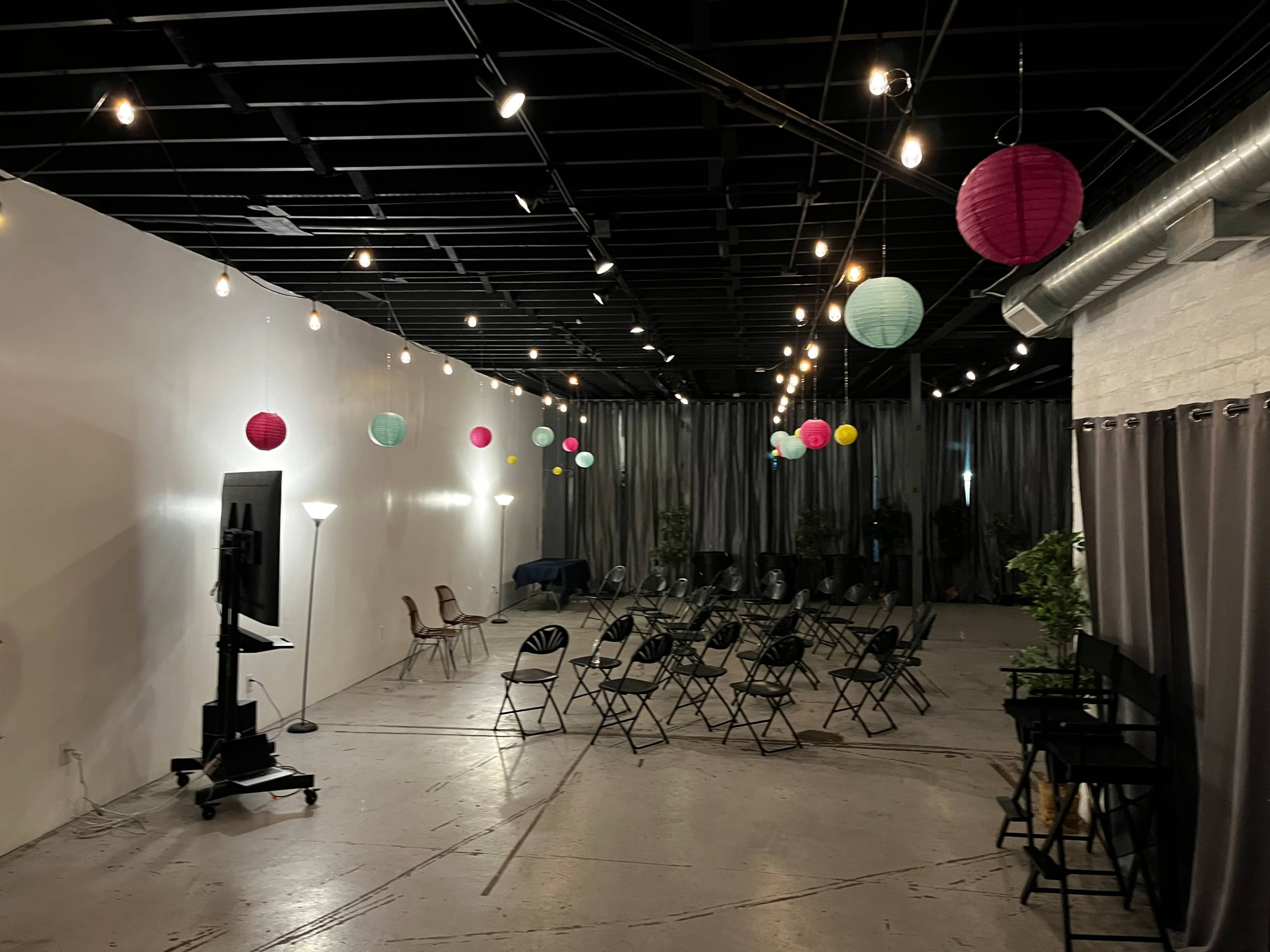 The image shows a spacious room with rows of chairs arranged for an event, decorated with colorful paper lanterns and string lights.