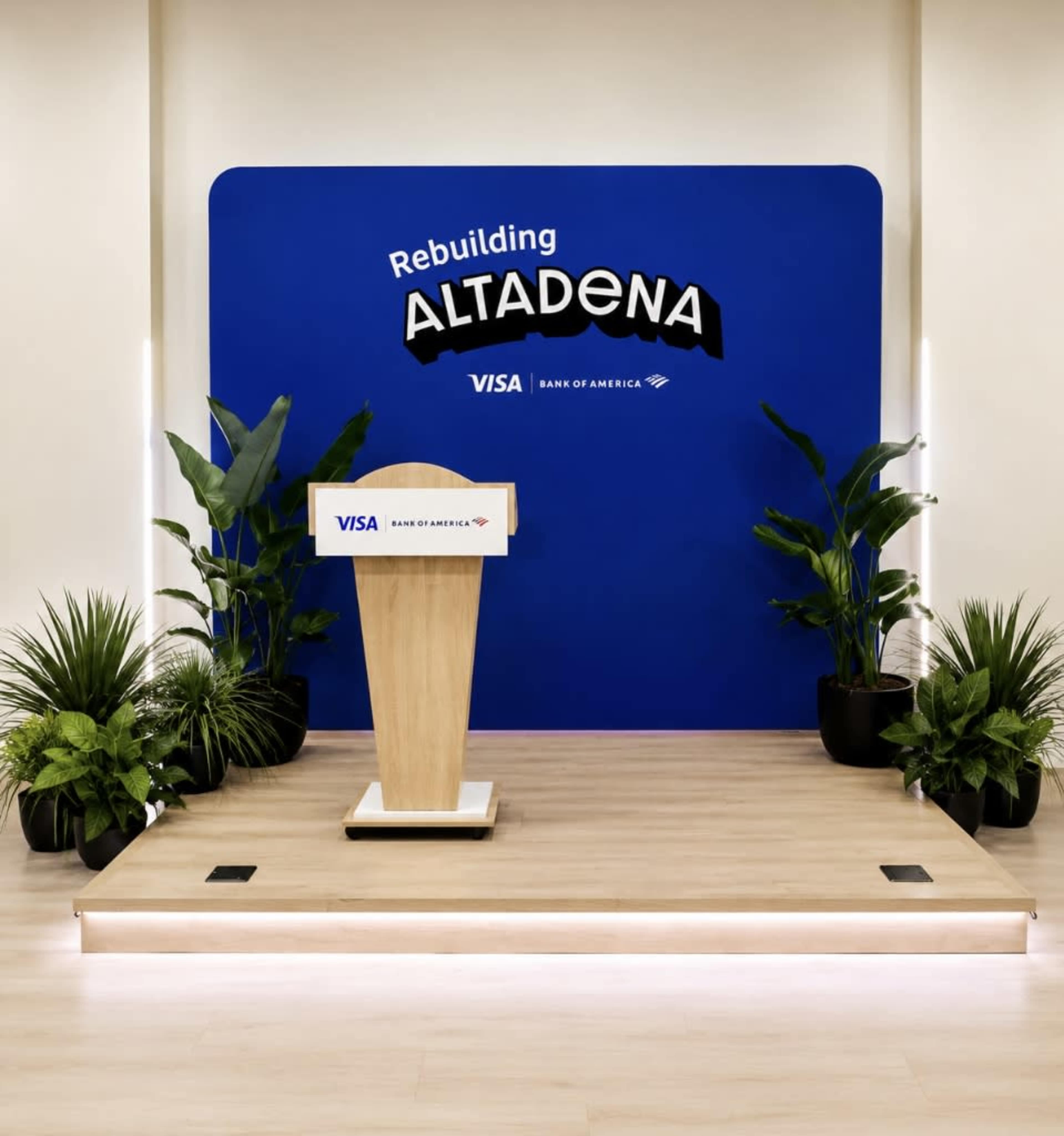 The image shows a podium set up in front of a blue backdrop displaying the text "Rebuilding ALTADENA" along with the logos of Visa and Bank of America, surrounded by potted plants.