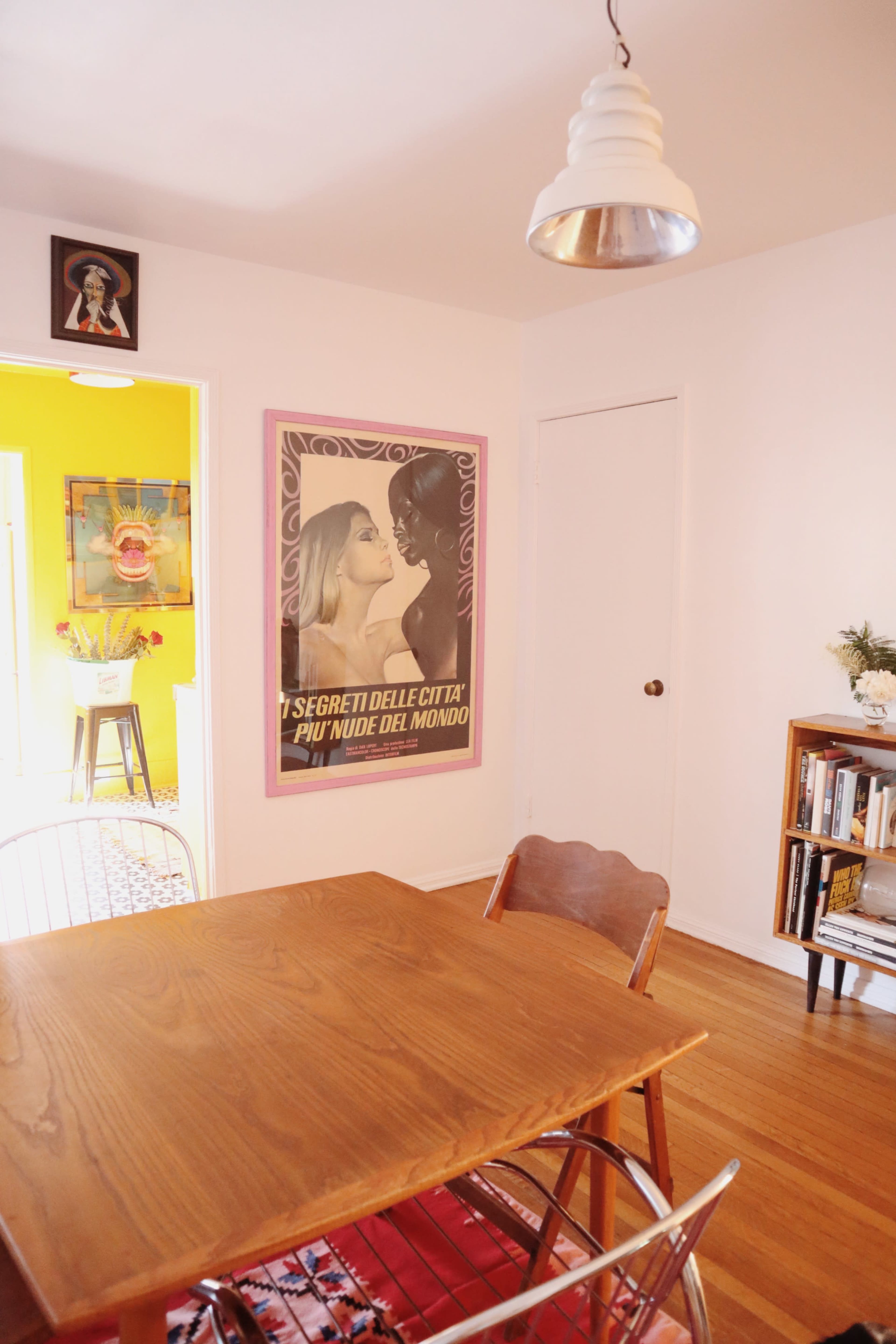 The image shows a dining area with a wooden table and chairs, featuring a large vintage-style poster on the wall and a doorway leading to a brightly colored room.