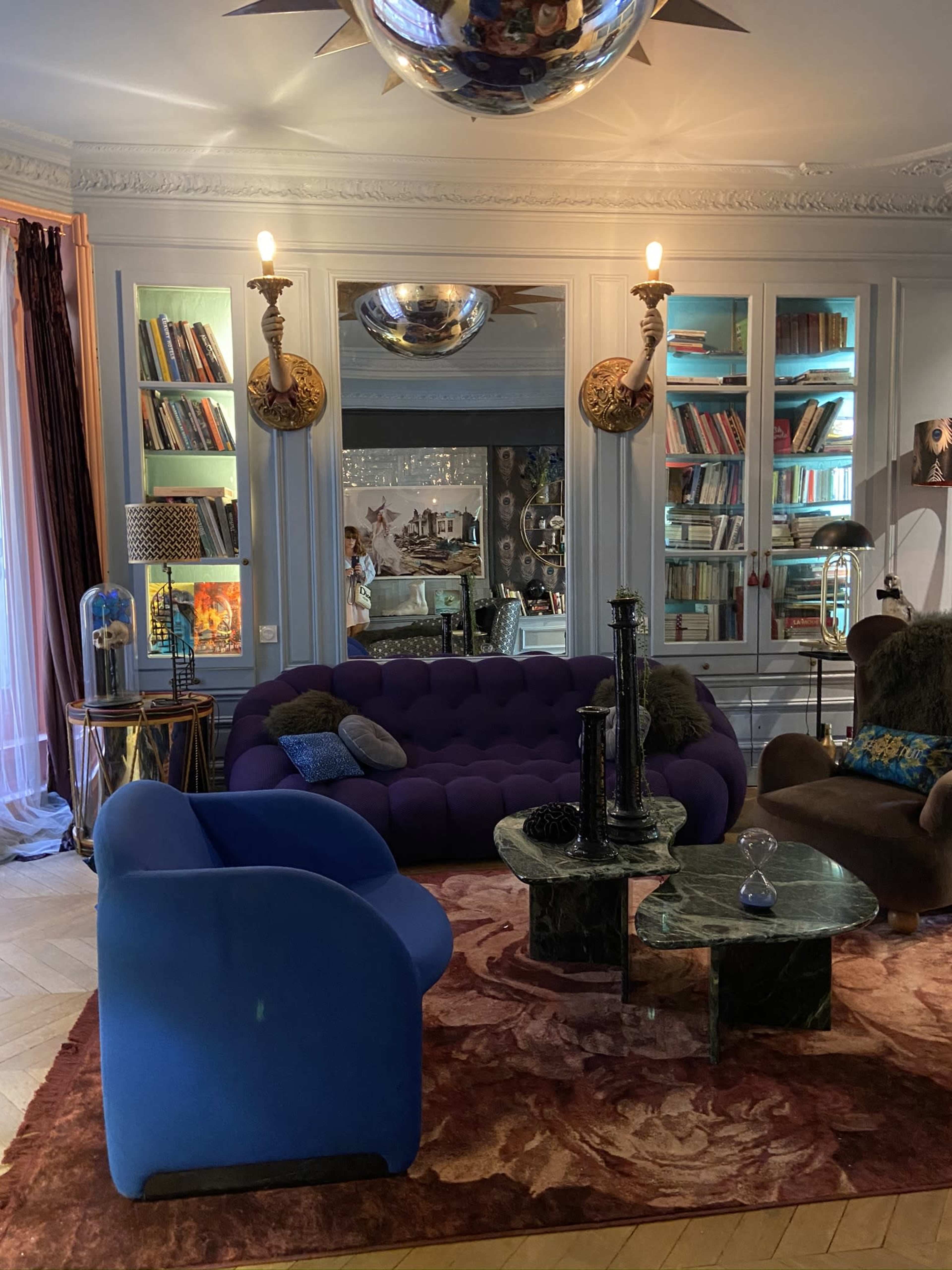 The image shows a stylish living room featuring a purple sofa, a blue armchair, a marble coffee table, and shelves filled with books.