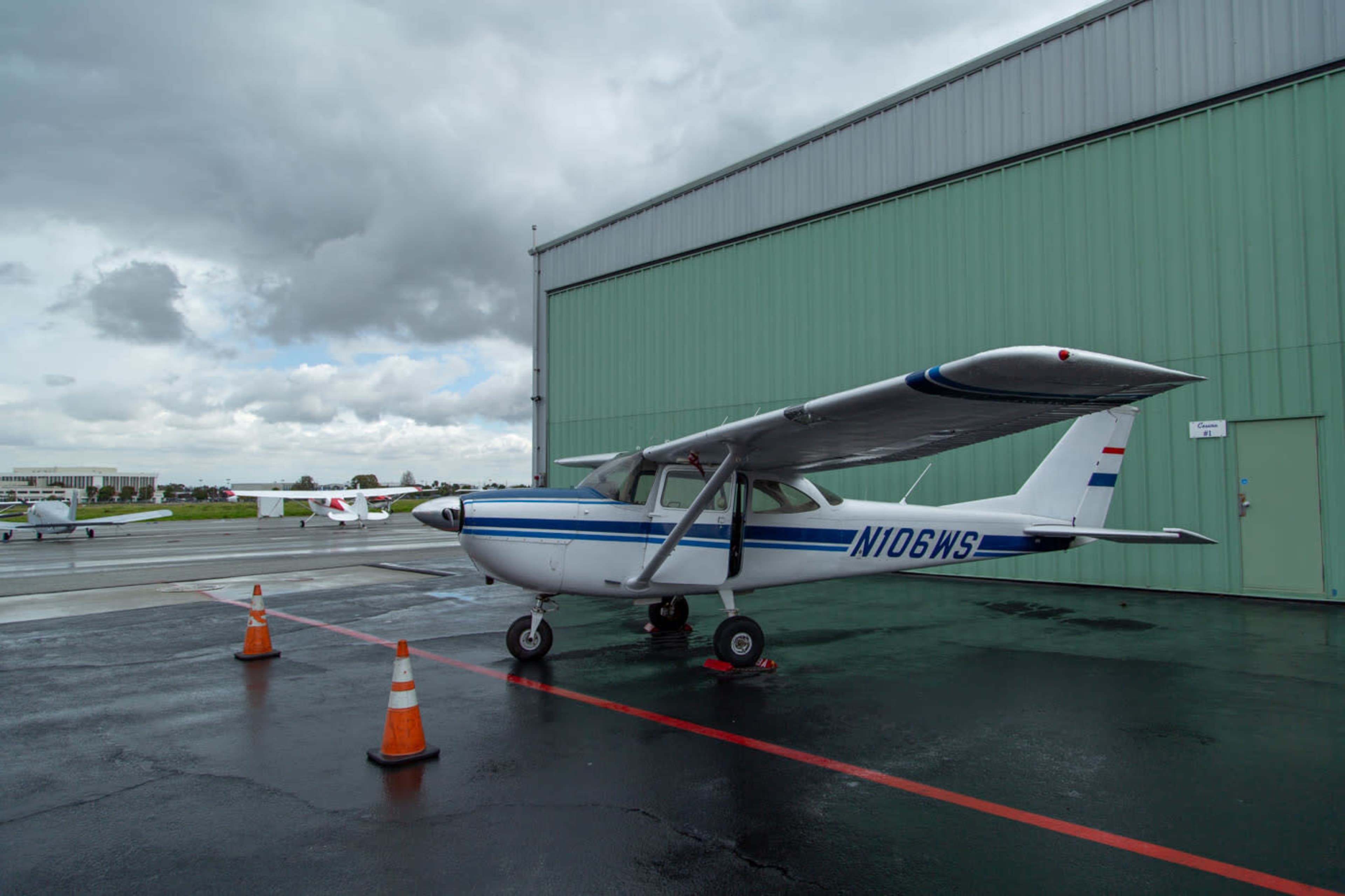 A single-engine Cessna aircraft is parked on a tarmac in front of a green hangar, with orange traffic cones marking its position.