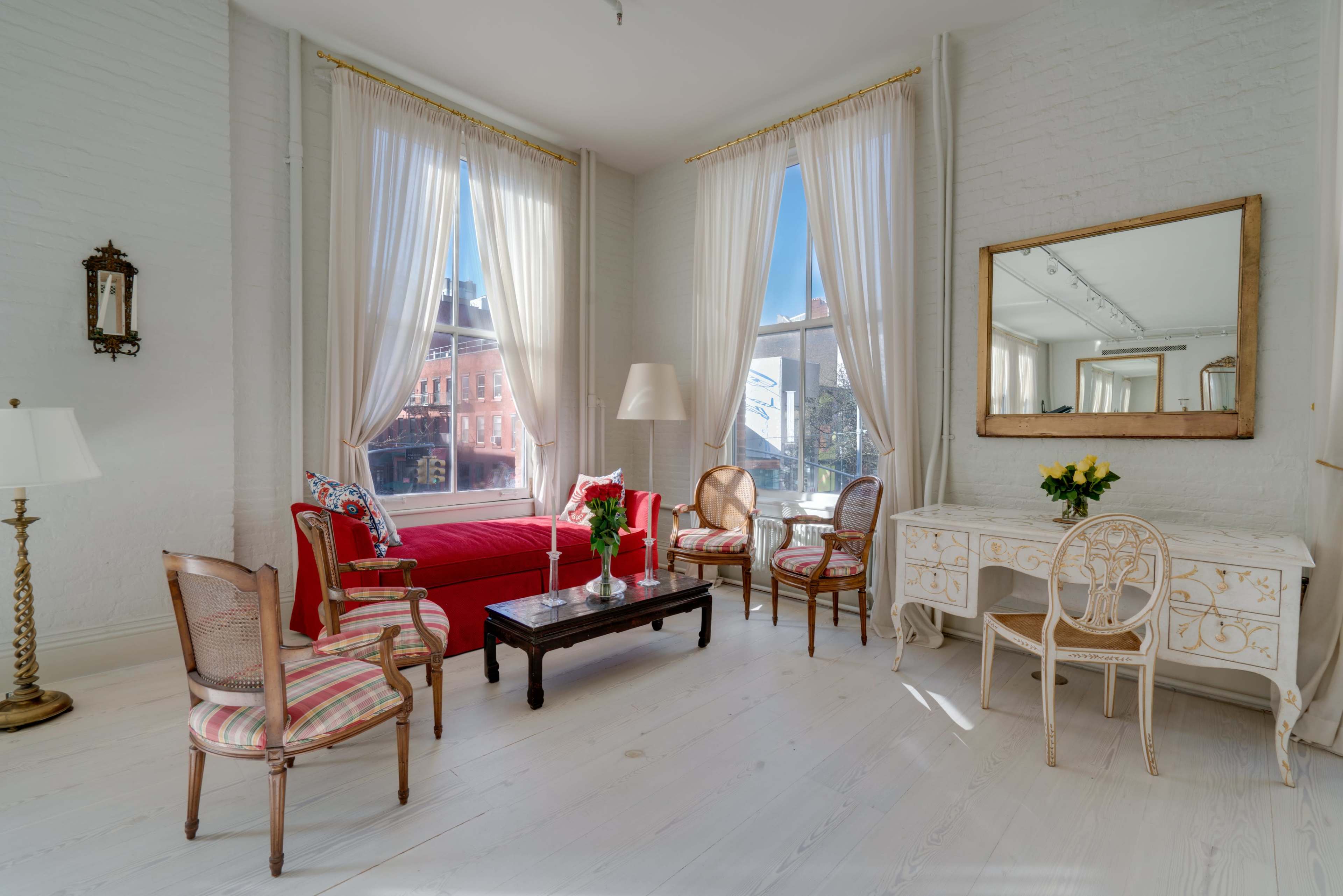 A bright and airy room features a red sofa, two chairs, a white wooden desk, and large windows with sheer curtains.