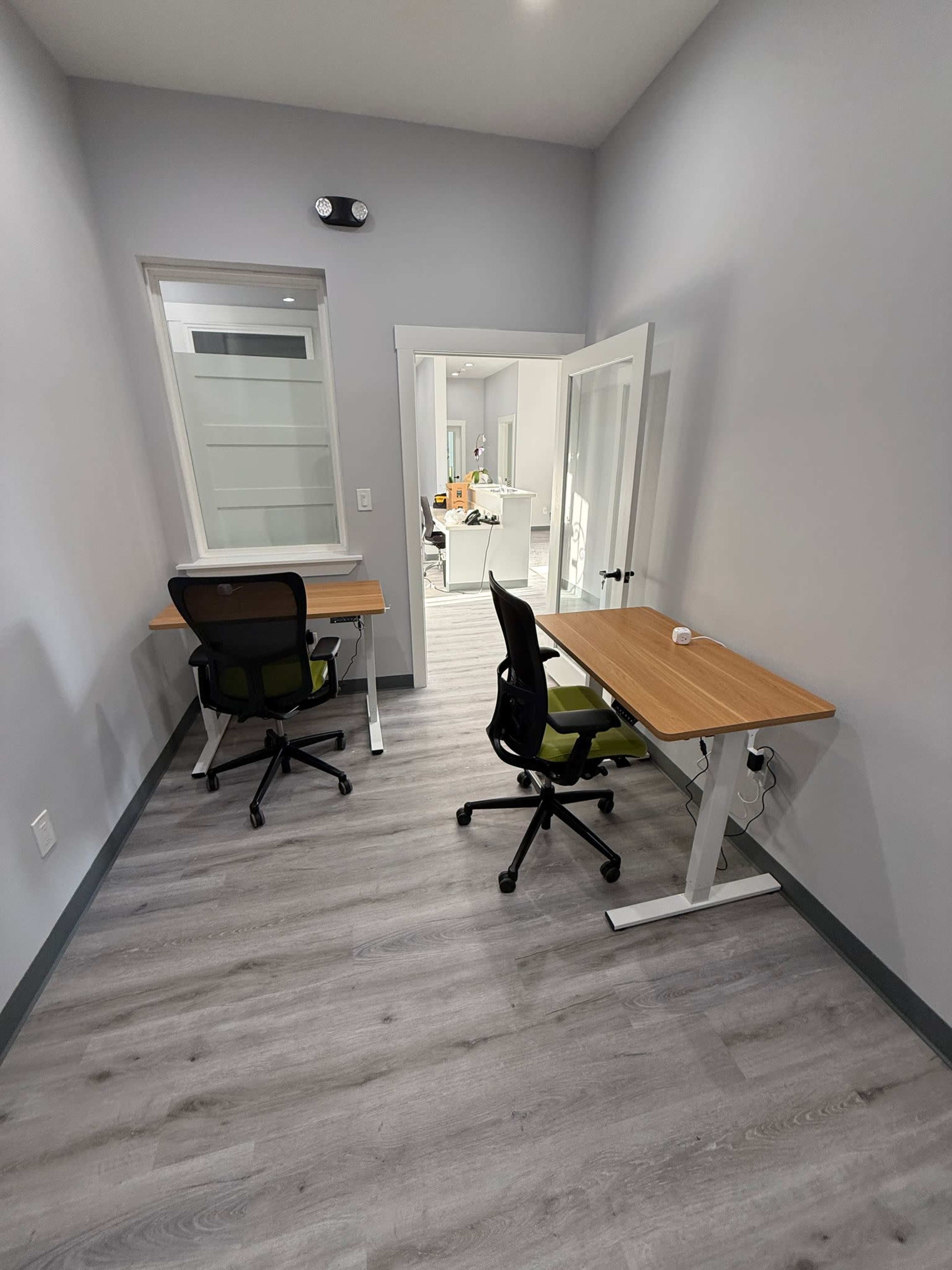 The image shows a small office space with two desks and ergonomic chairs positioned against a wall, with a door leading to another room in the background.